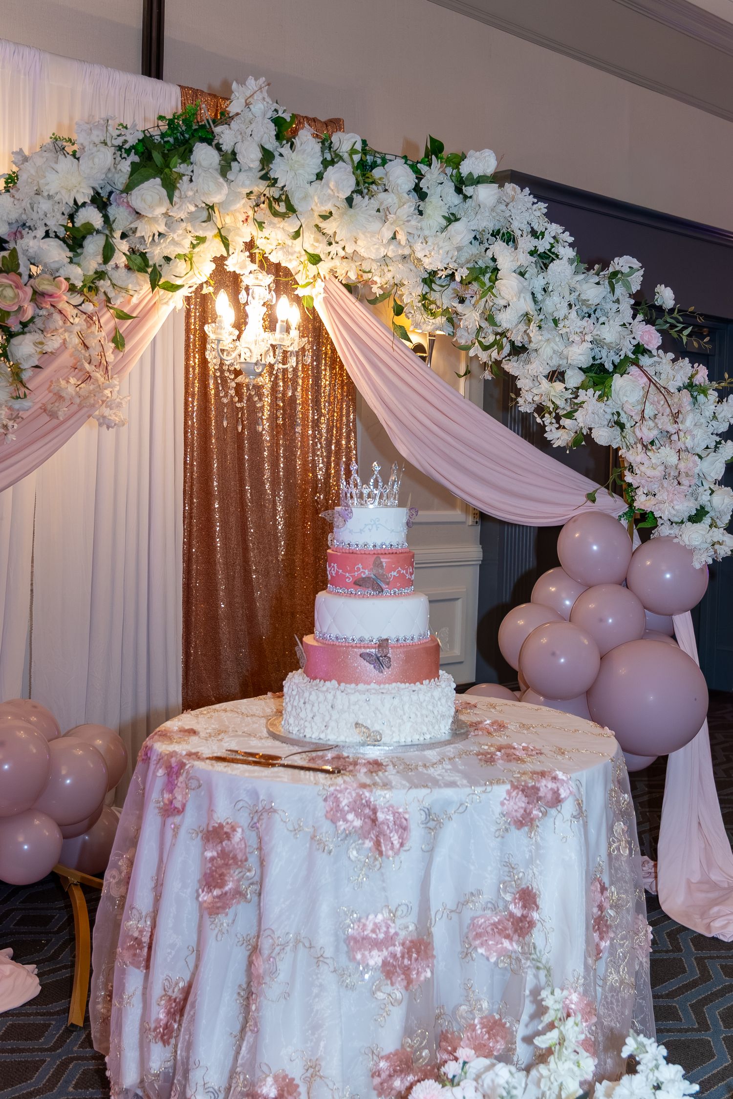 Elegant multi-tier wedding cake with crown topper on floral table under white flower arch with pink balloons. Sweet 16/Quinceañera