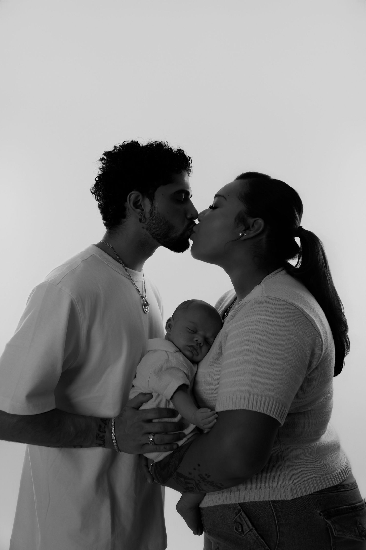 Black and white photo of a couple/parents kissing while holding their newborn baby in a studio setting. Newborn and family photoshoot.