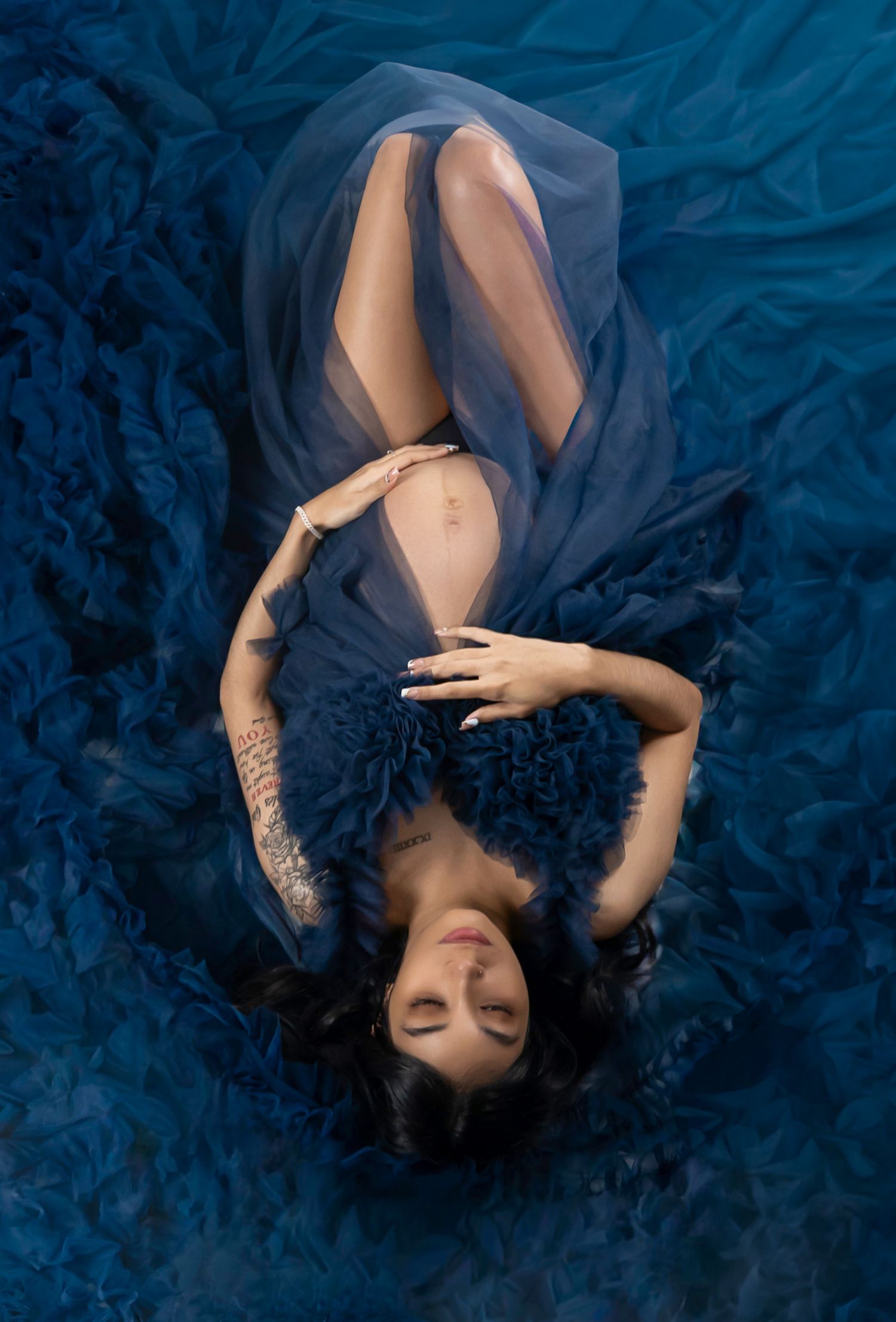 A woman in a flowing blue ruffled gown lies gracefully on a blue fabric backdrop, viewed from above.