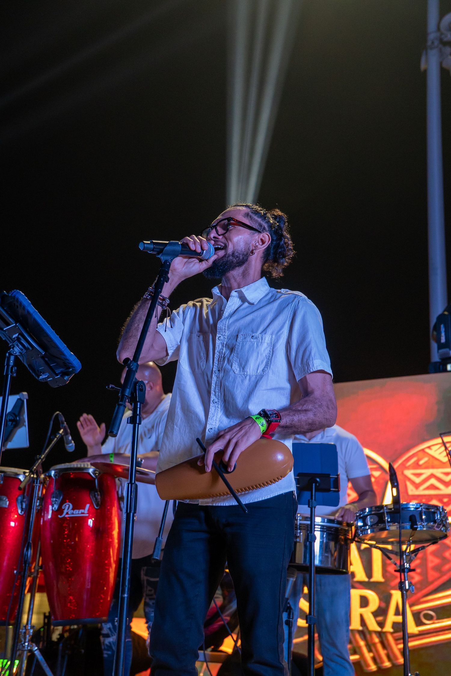 Male singer performing on stage with microphone, wearing white shirt, with conga drums and stage lighting behind him.