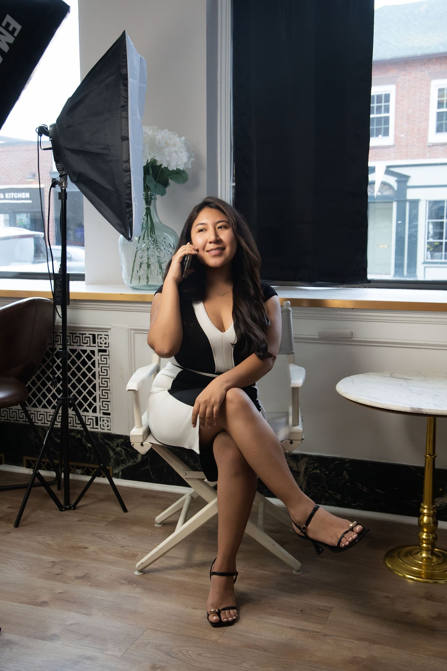 Young woman in white and black dress sits in director's chair by window with studio lighting equipment.