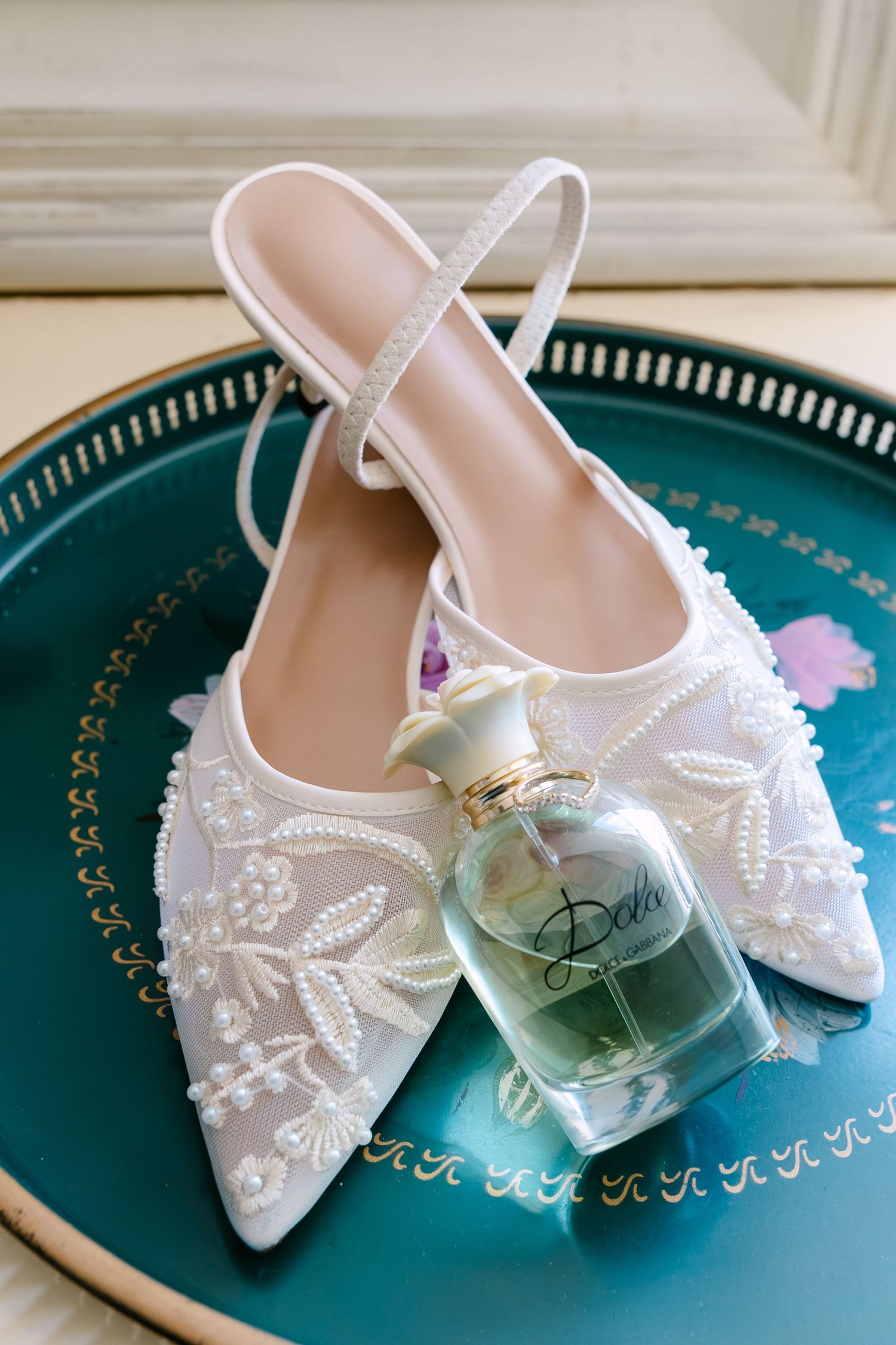 White beaded bridal heels with floral embroidery beside a Dolce perfume bottle on a teal decorative tray.
