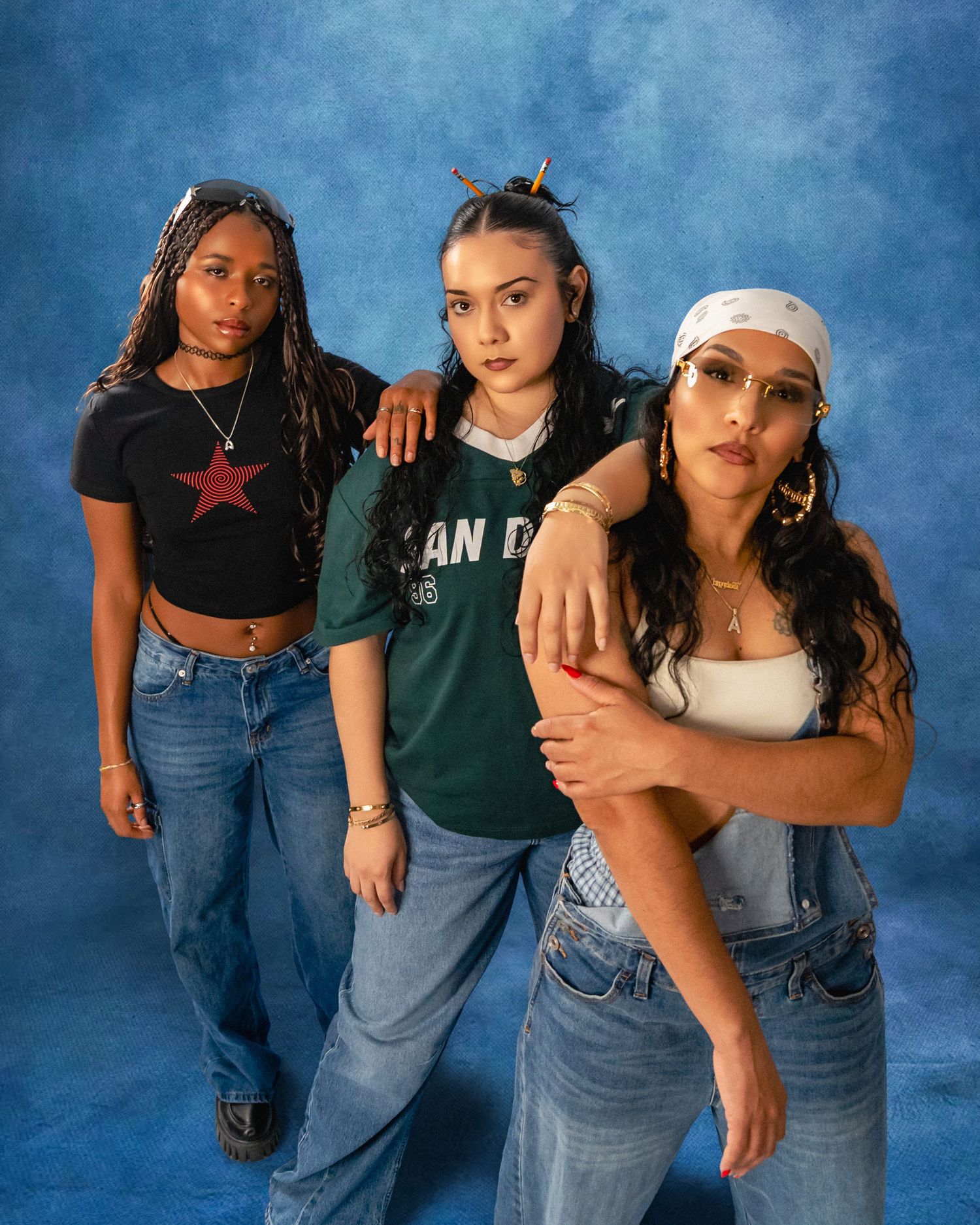 Three young women in Y2K-inspired denim and graphic tees pose closely against a blue studio backdrop, one wearing a white bandana.