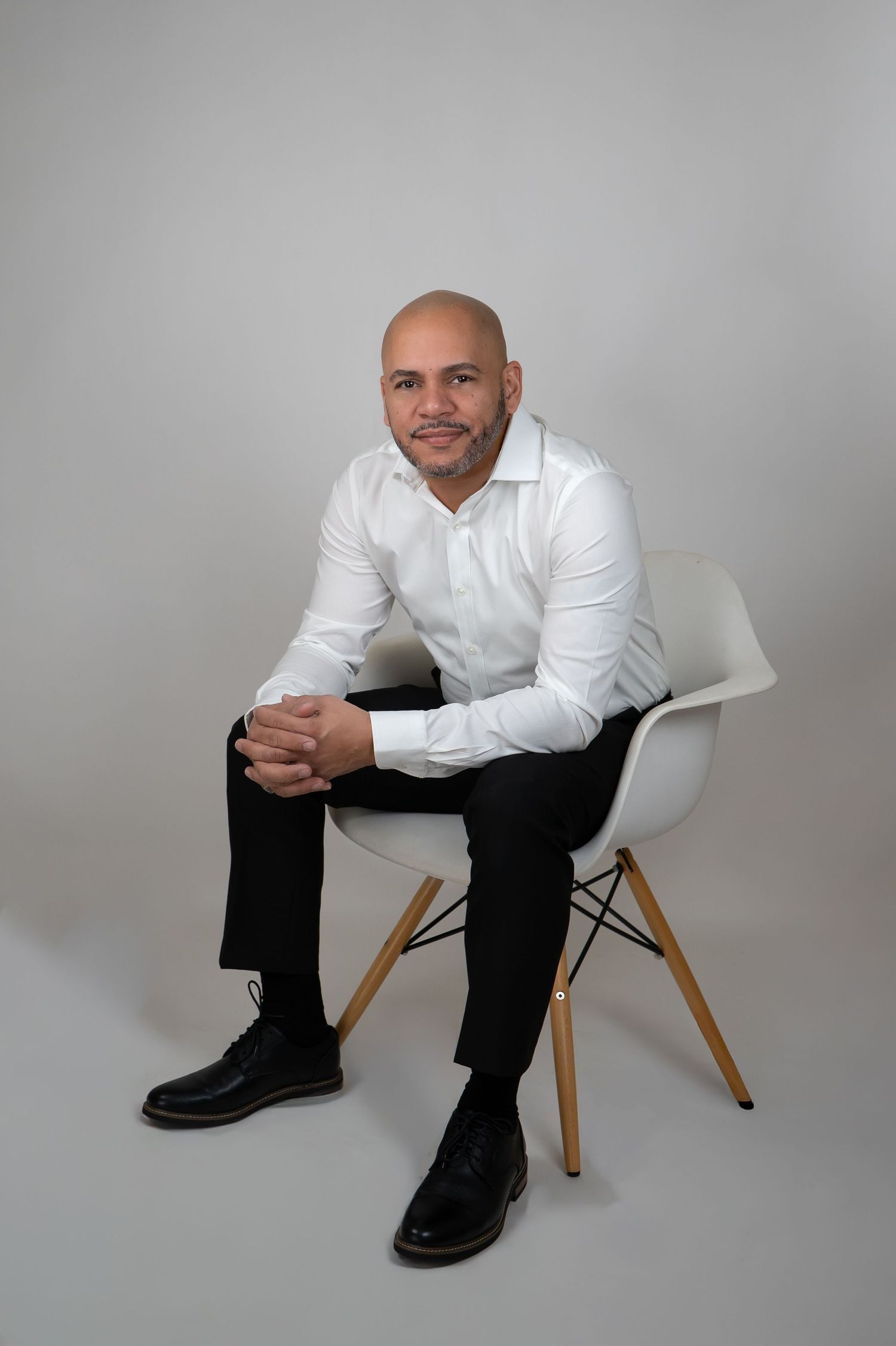 Bald man in white shirt and black pants sits relaxed on modern white chair.