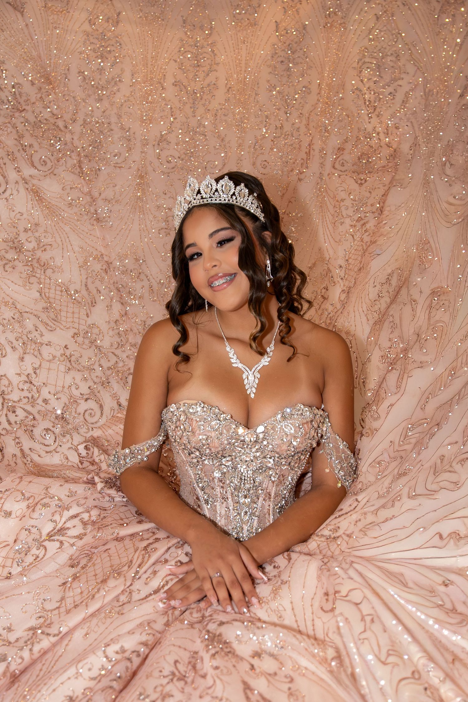 Young woman in a beaded rose gold off-shoulder quinceañera gown and crystal tiara poses elegantly. Sweet 16/Quinceañera