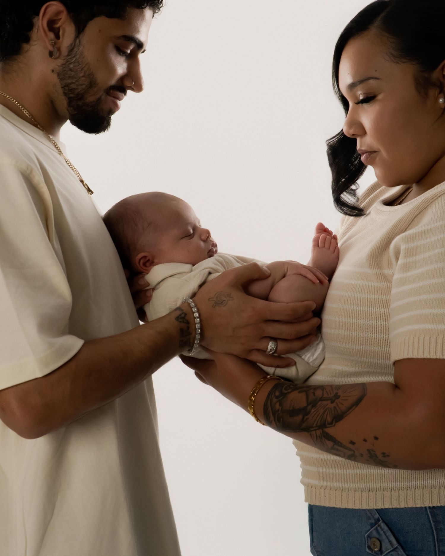 A couple tenderly holds their newborn baby together, both gazing lovingly at the infant in a soft, neutral studio setting. Newborn photoshoo