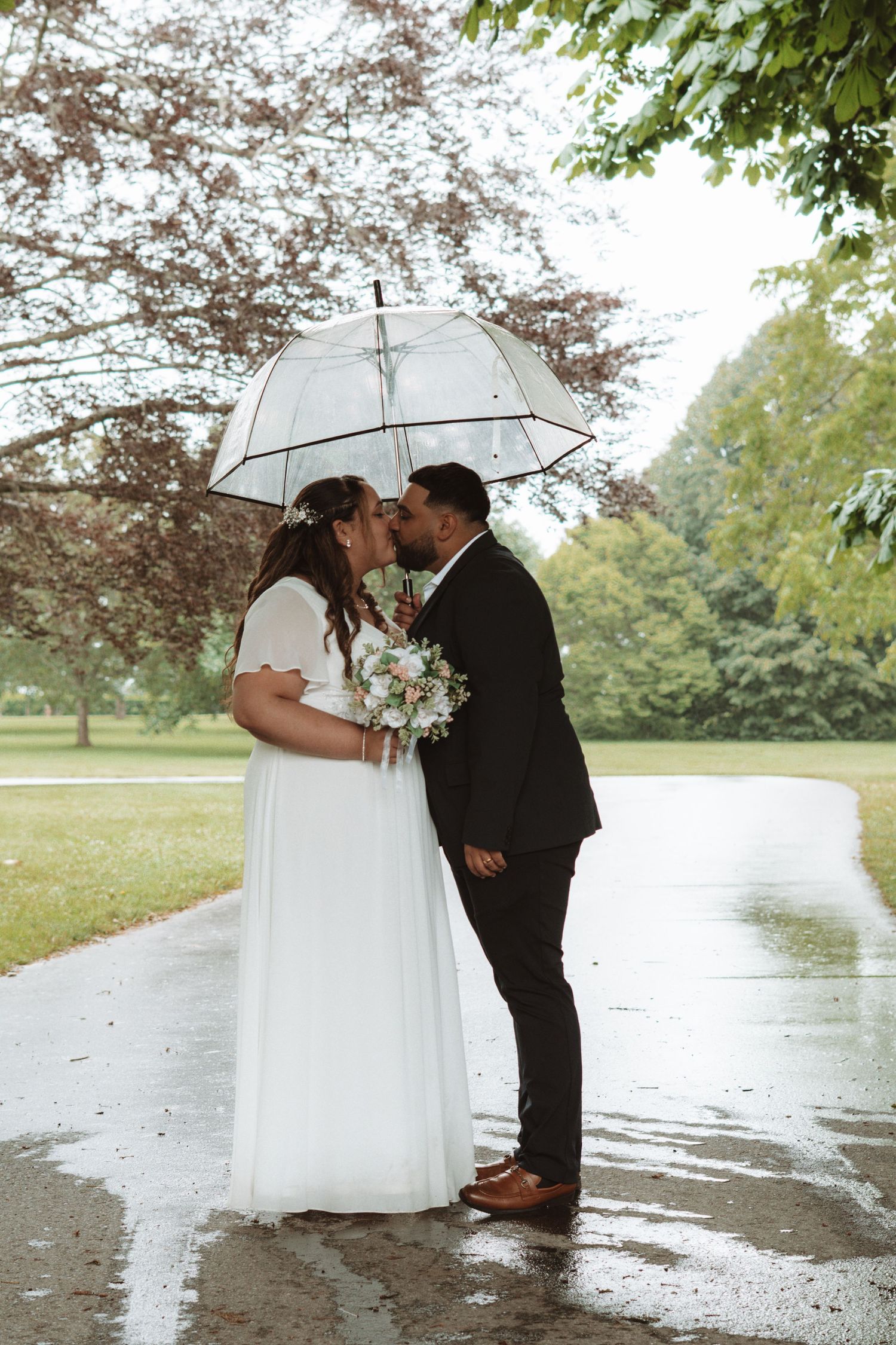 Bride and groom share a romantic kiss under a clear bubble umbrella on a rainy wedding day in a lush park.
