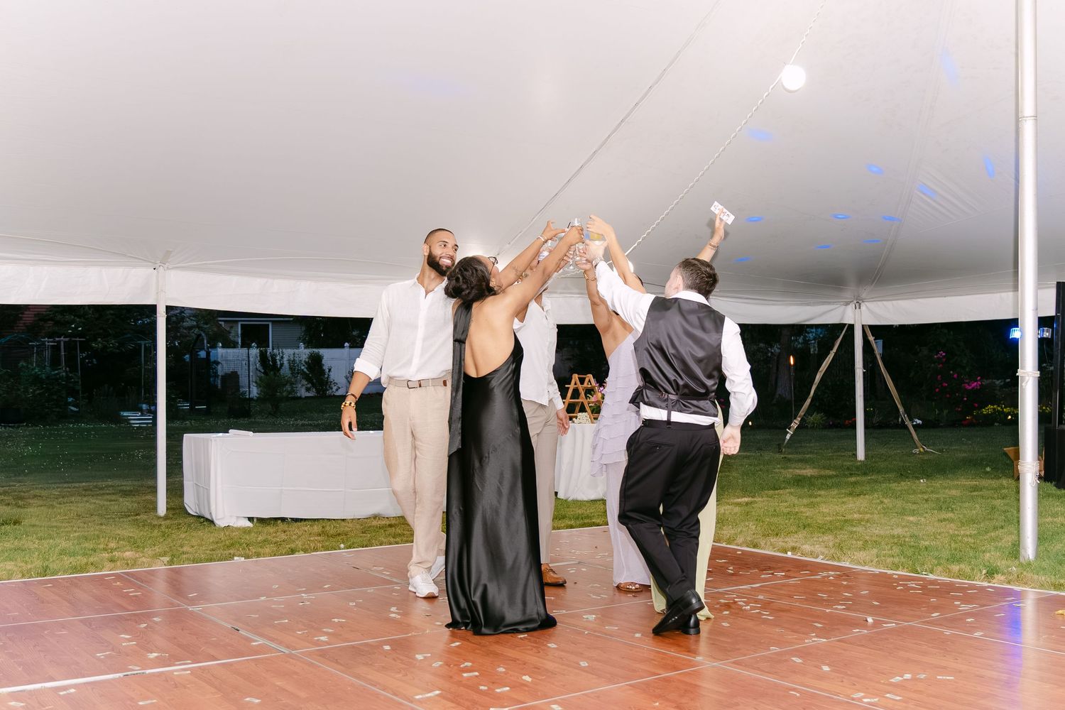 Wedding guests dancing together under a white tent at an outdoor evening reception on a wooden dance floor.