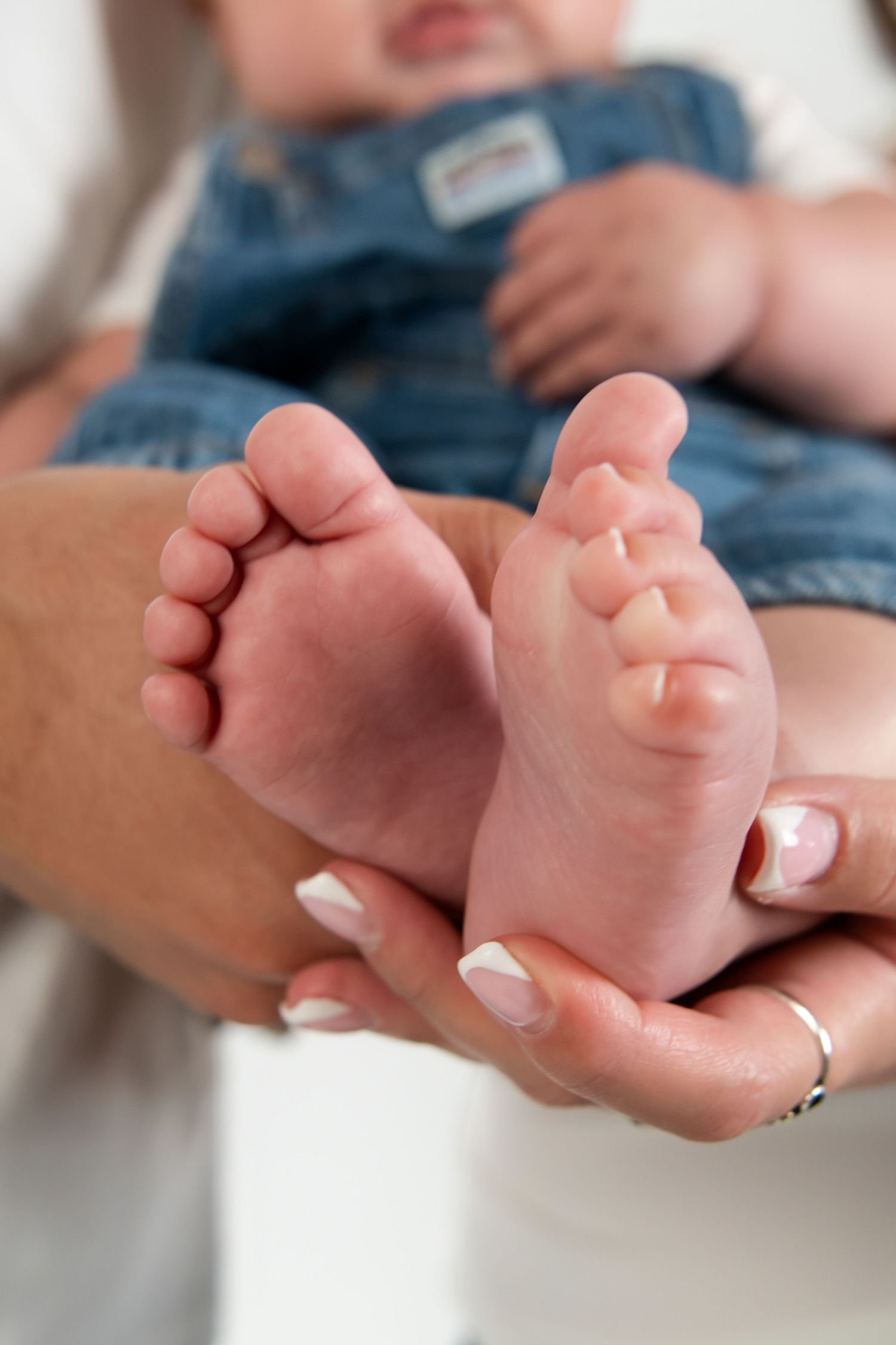 Adult hands with french manicure gently cradle the tiny bare feet of a baby wearing denim overalls.