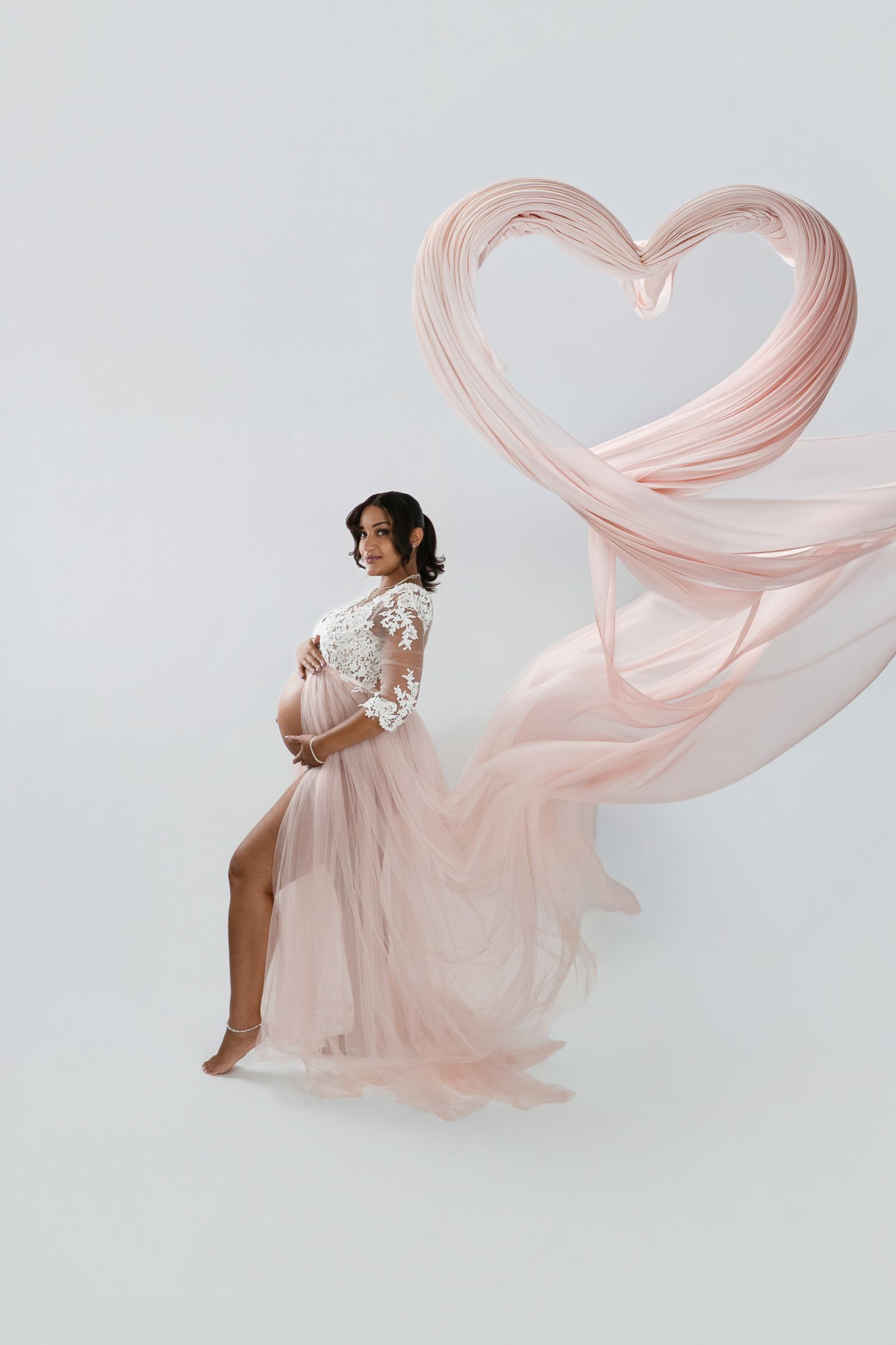 Pregnant woman in a blush pink lace and tulle maternity gown cradling her belly as flowing fabric forms a heart shape in a studio.