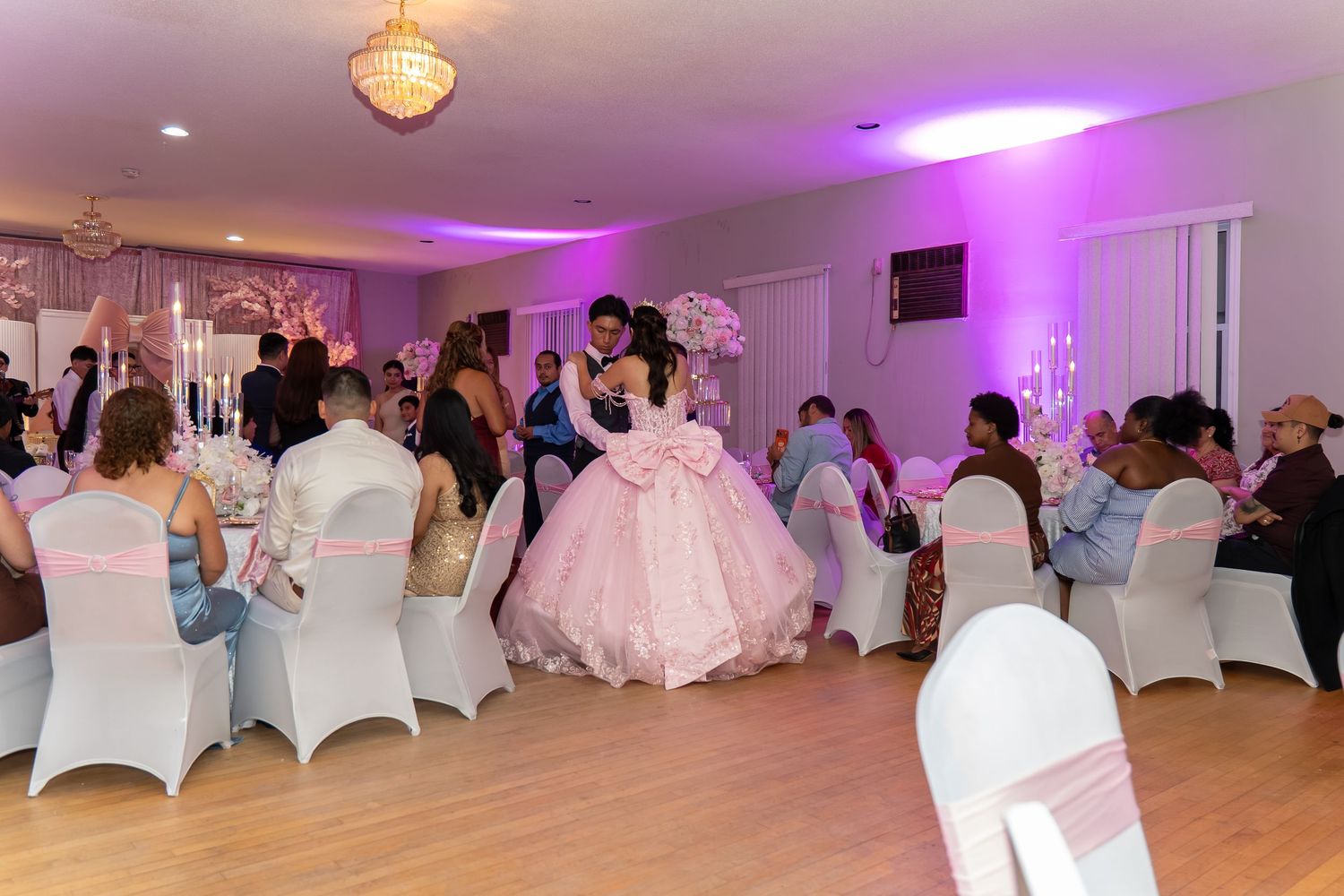 Young woman in pink ball gown dances with partner at elegant quinceañera celebration with purple lighting.