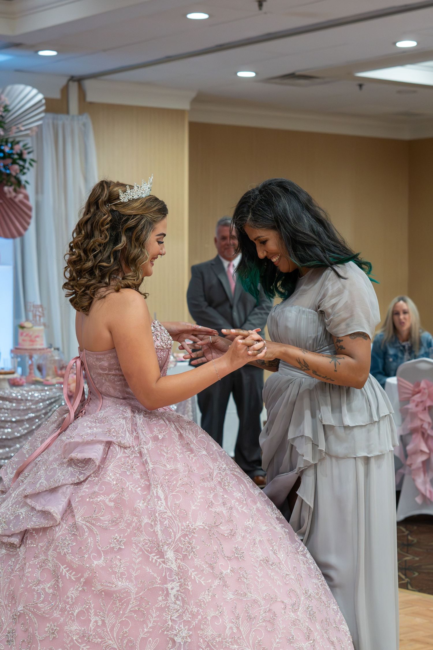 A young woman in a pink quinceañera gown and tiara receives a ring from an older woman in a gray dress at her celebration. Sweet 16!