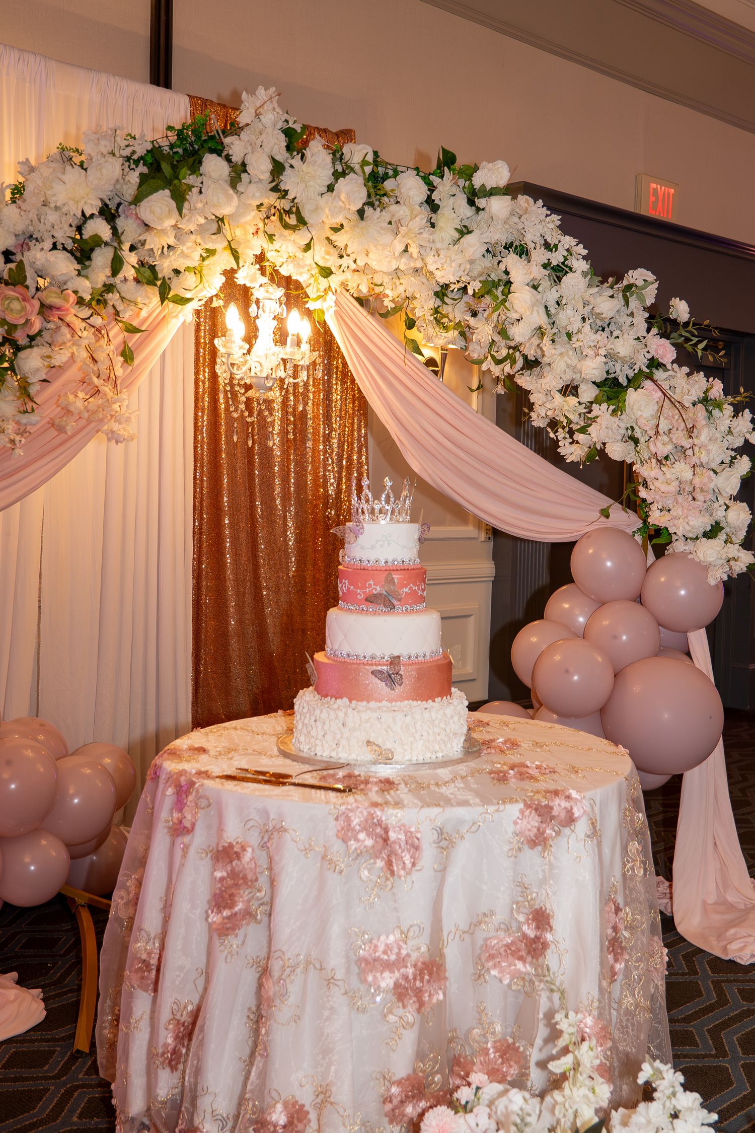 Elegant multi-tier pink and white wedding cake on floral tablecloth under a floral arch with balloons. Sweet 16/Quinceañera