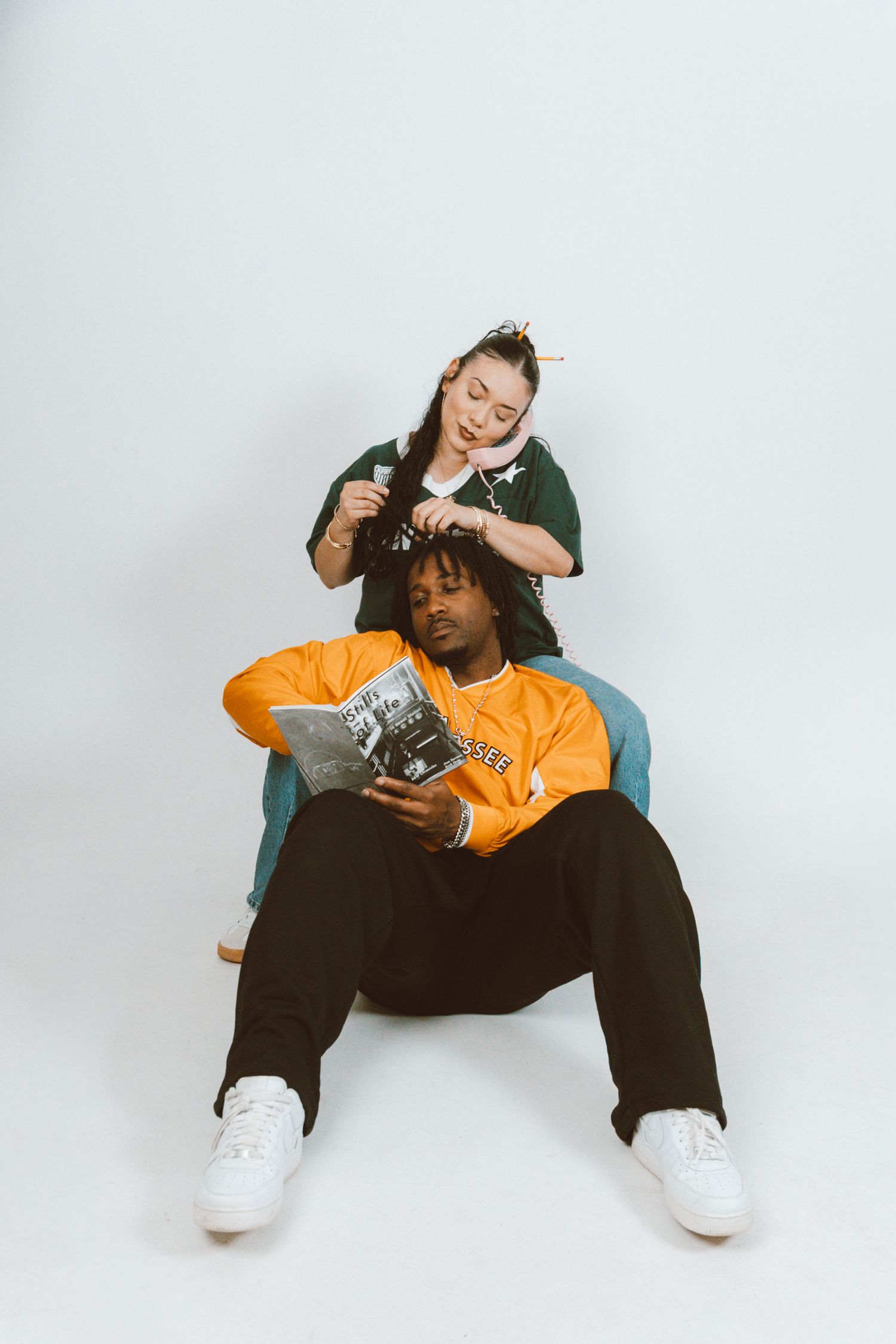 Man in an orange jersey and black pants reads a magazine on the floor while a woman in a green shirt sits behind him braiding his hair.