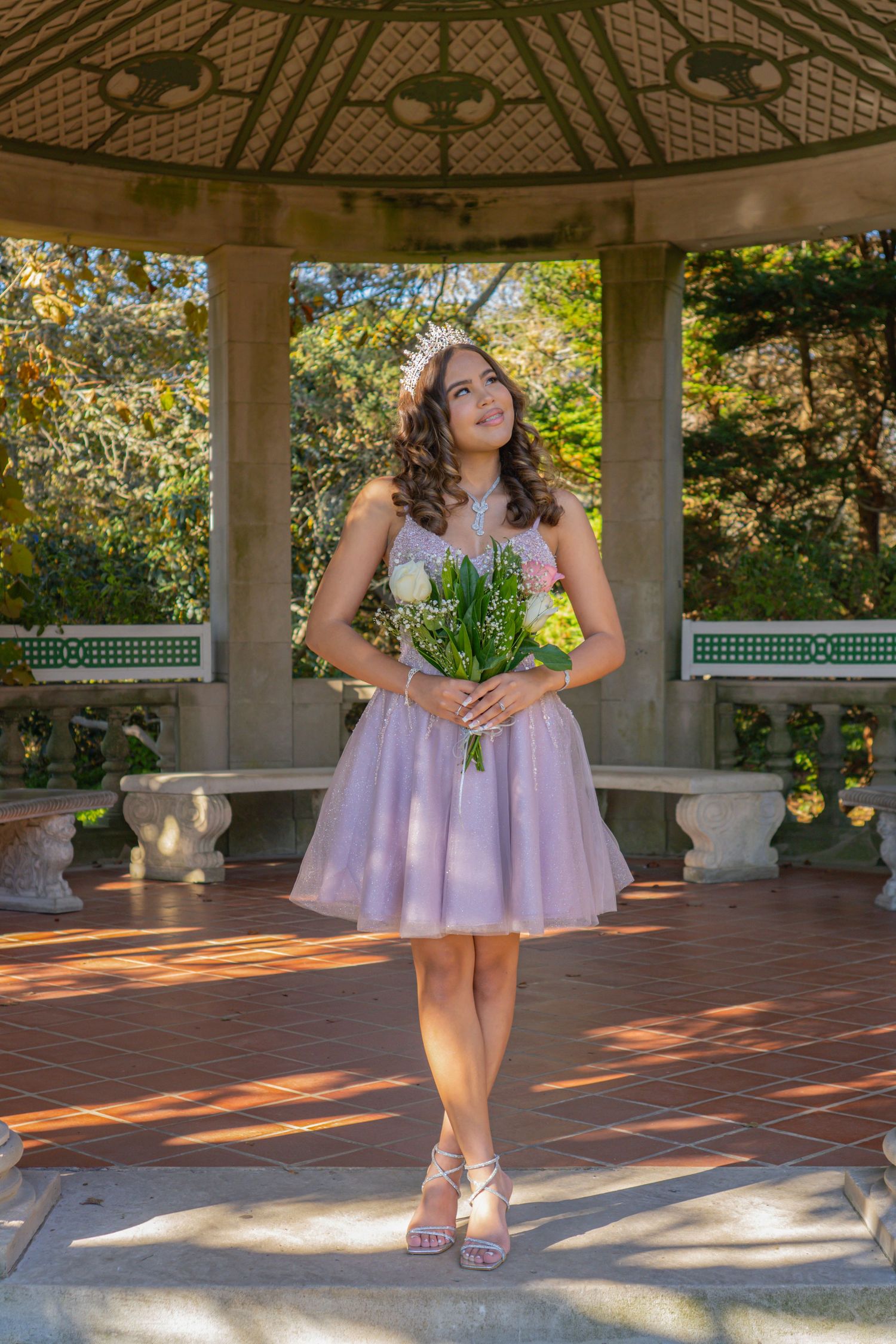 Young woman in lavender tulle dress holds white flowers in a sunlit garden gazebo.
