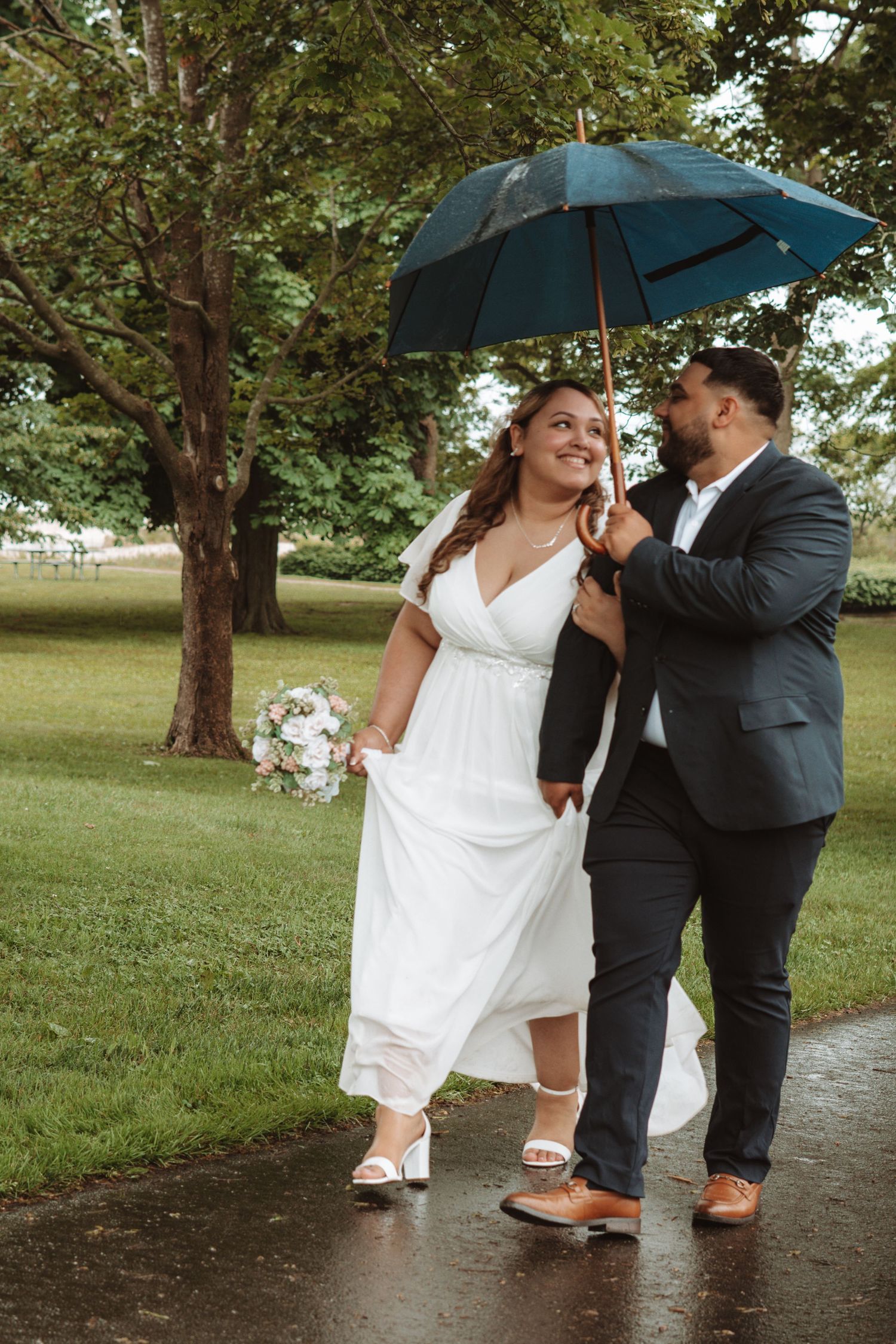 Bride holding bouquet and groom walking together under a teal umbrella on a rainy wedding day in the park.