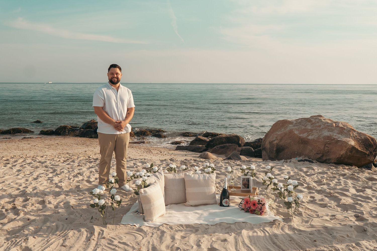 Man stands on sandy beach beside romantic picnic setup with flowers, cushions, and wine, ocean and rocks in background.