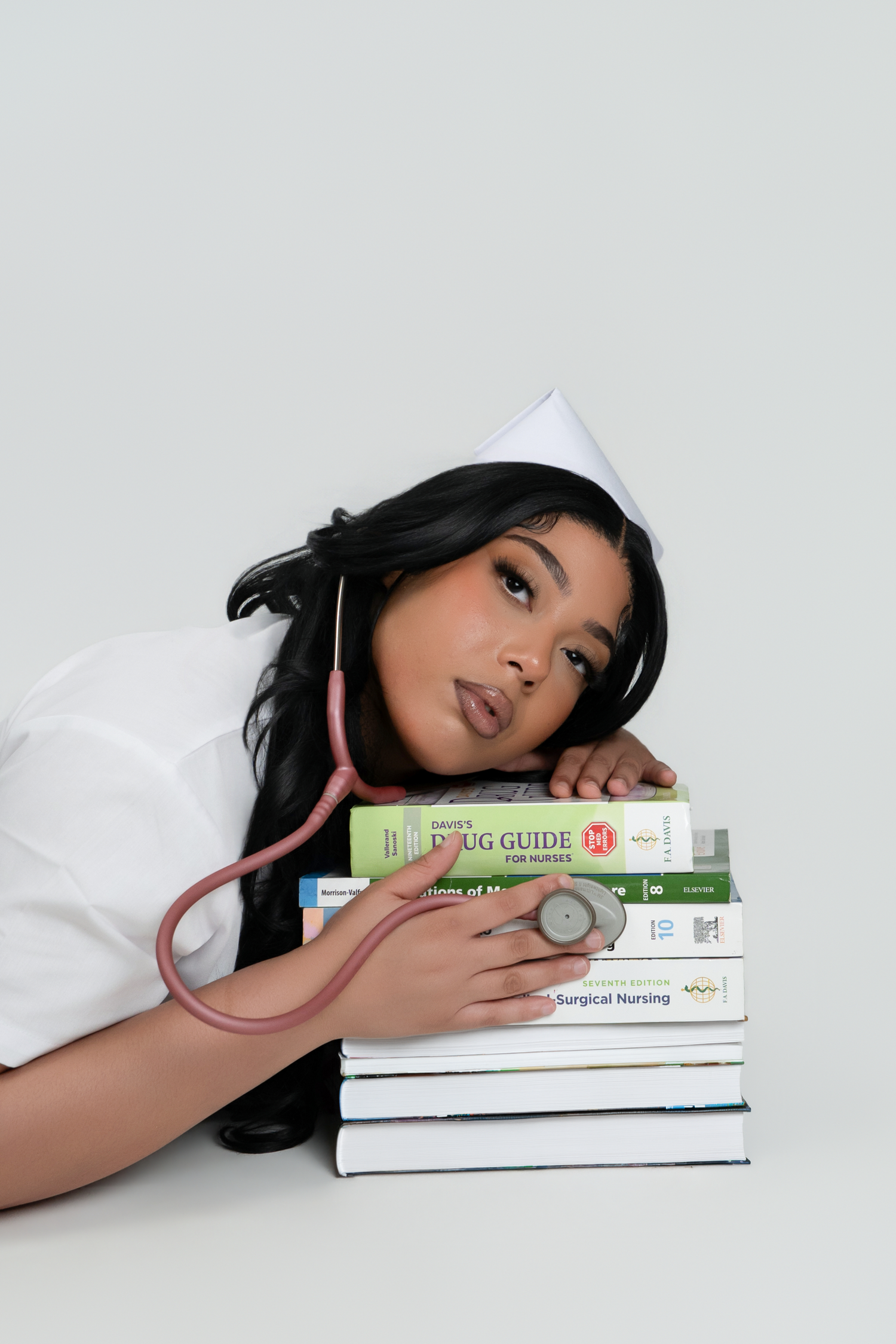 A nursing student in white uniform and cap rests her head on a stack of medical textbooks with a pink stethoscope.