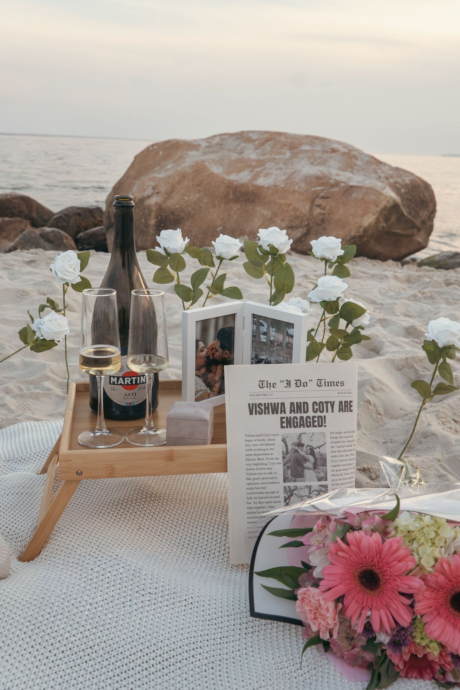 Romantic beach engagement setup with Martini, white roses, and a newspaper reading Vishwa and Coty Are Engaged.