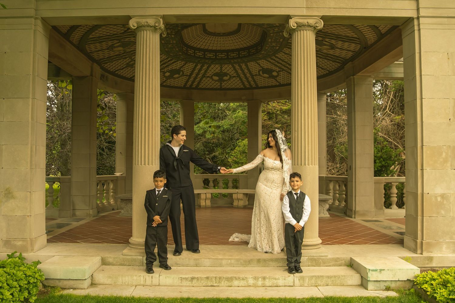 Wedding couple holding hands under elegant gazebo with two young boys in formal attire at outdoor garden venue.