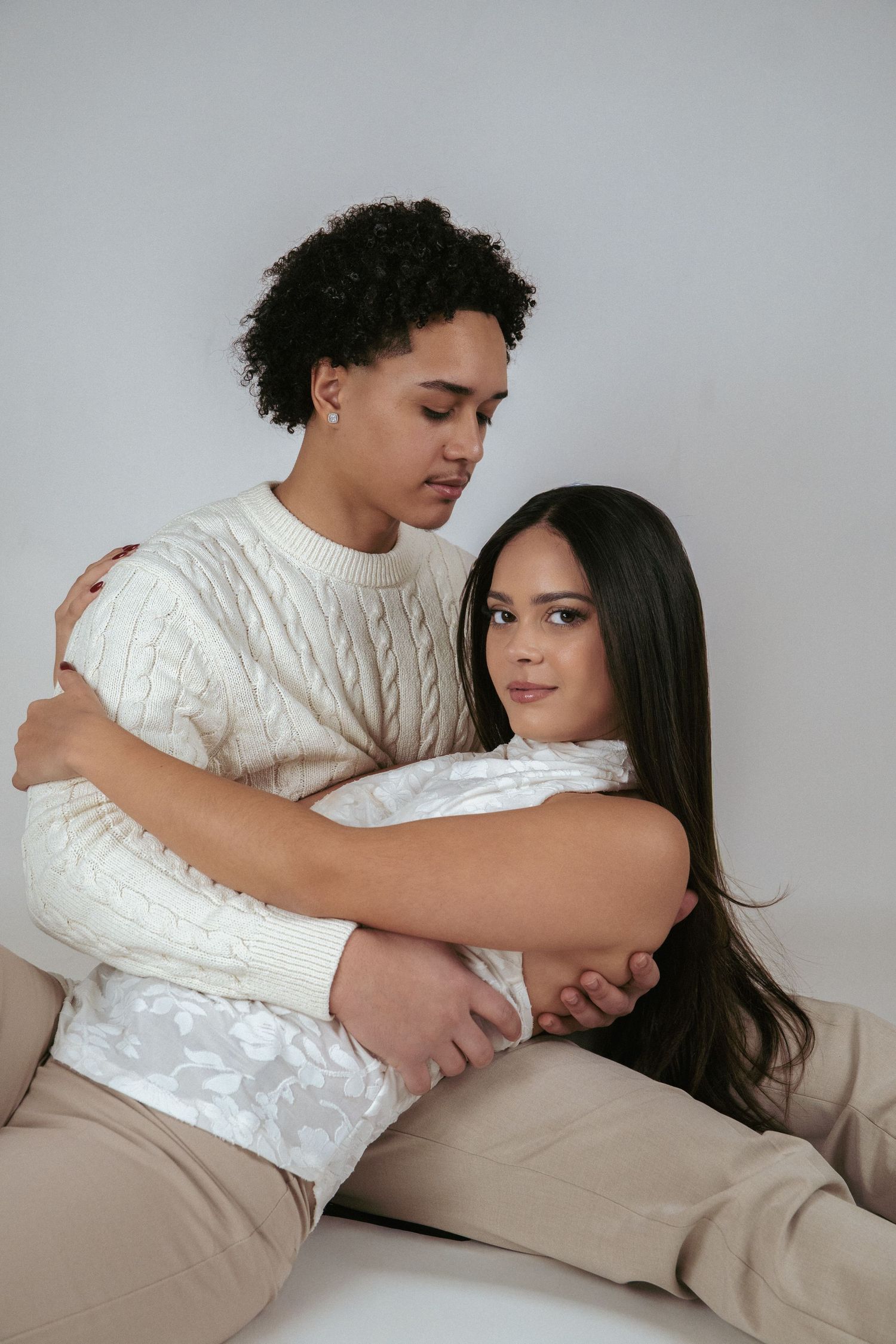 A young couple embracing warmly, the man in a cream cable-knit sweater holding the woman close against a white background.