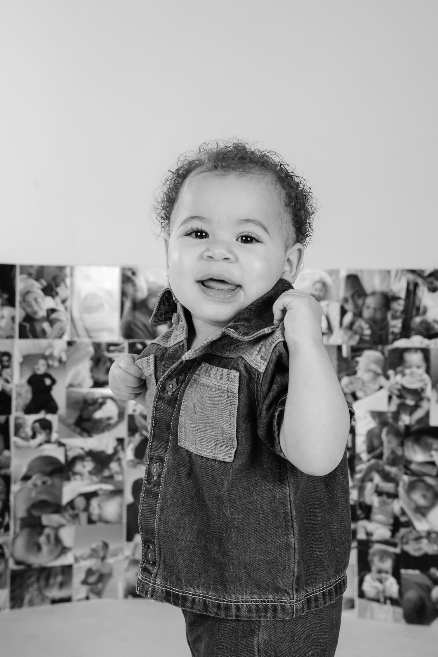 Black and white portrait of a smiling curly-haired toddler in a denim shirt standing in front of a photo collage wall.
