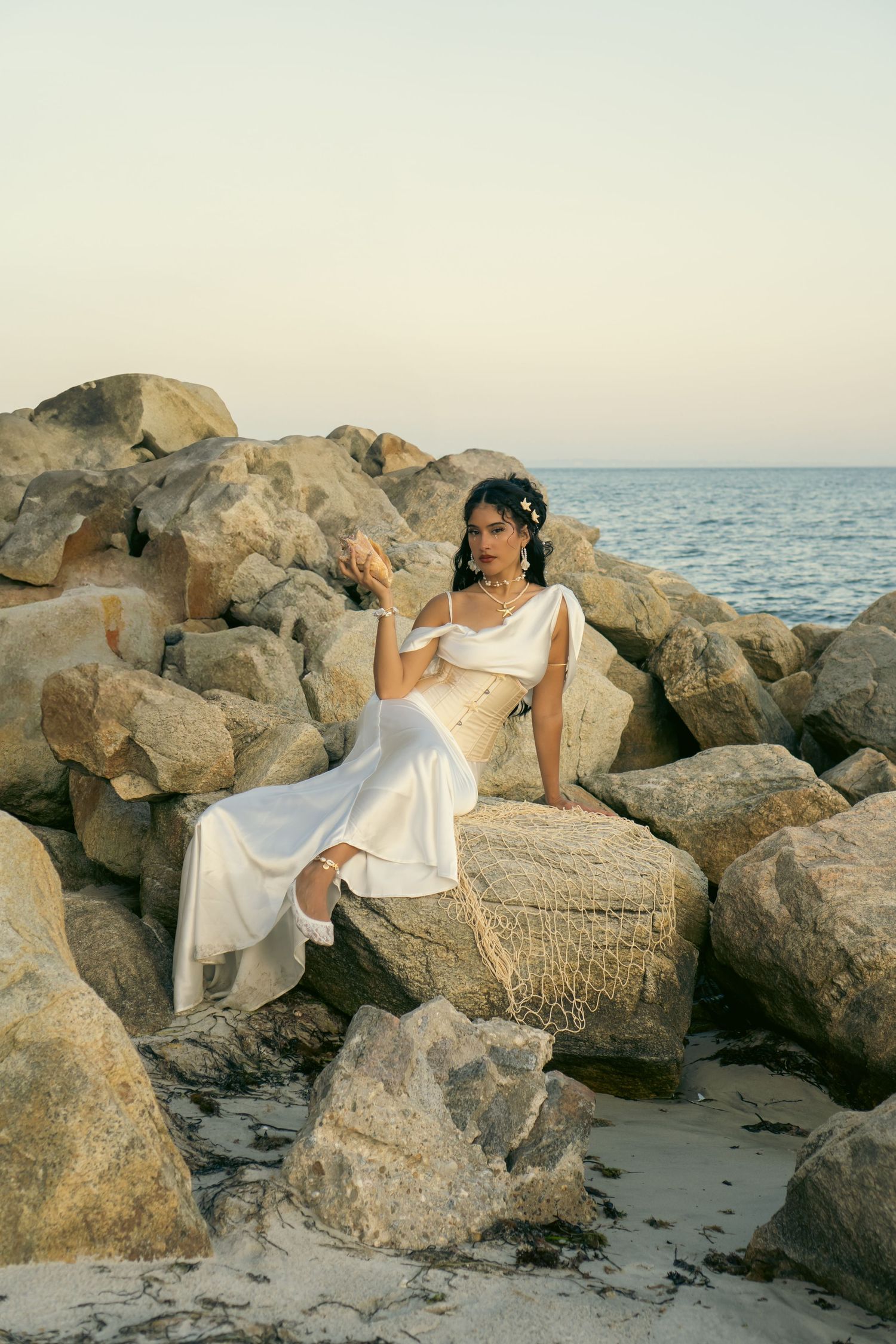 Woman in elegant white satin gown sitting gracefully on coastal rocks at sunset by the ocean.
