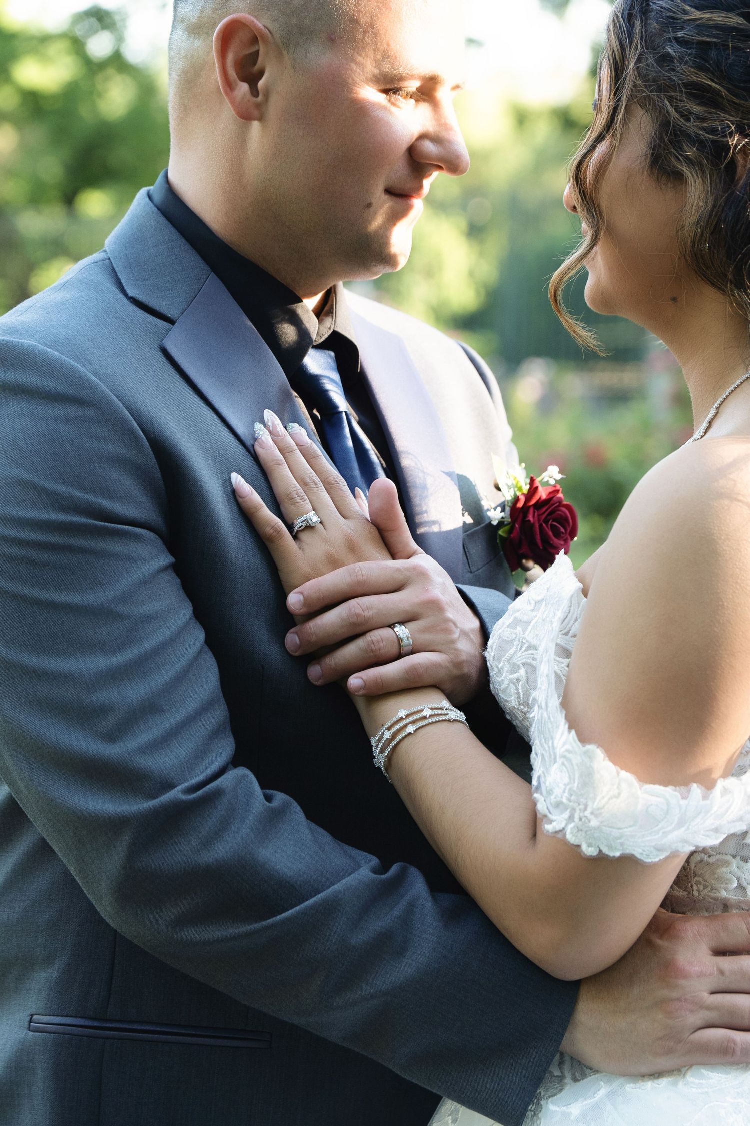 Bride and groom in wedding attire embracing outdoors, showing wedding rings and floral boutonniere.