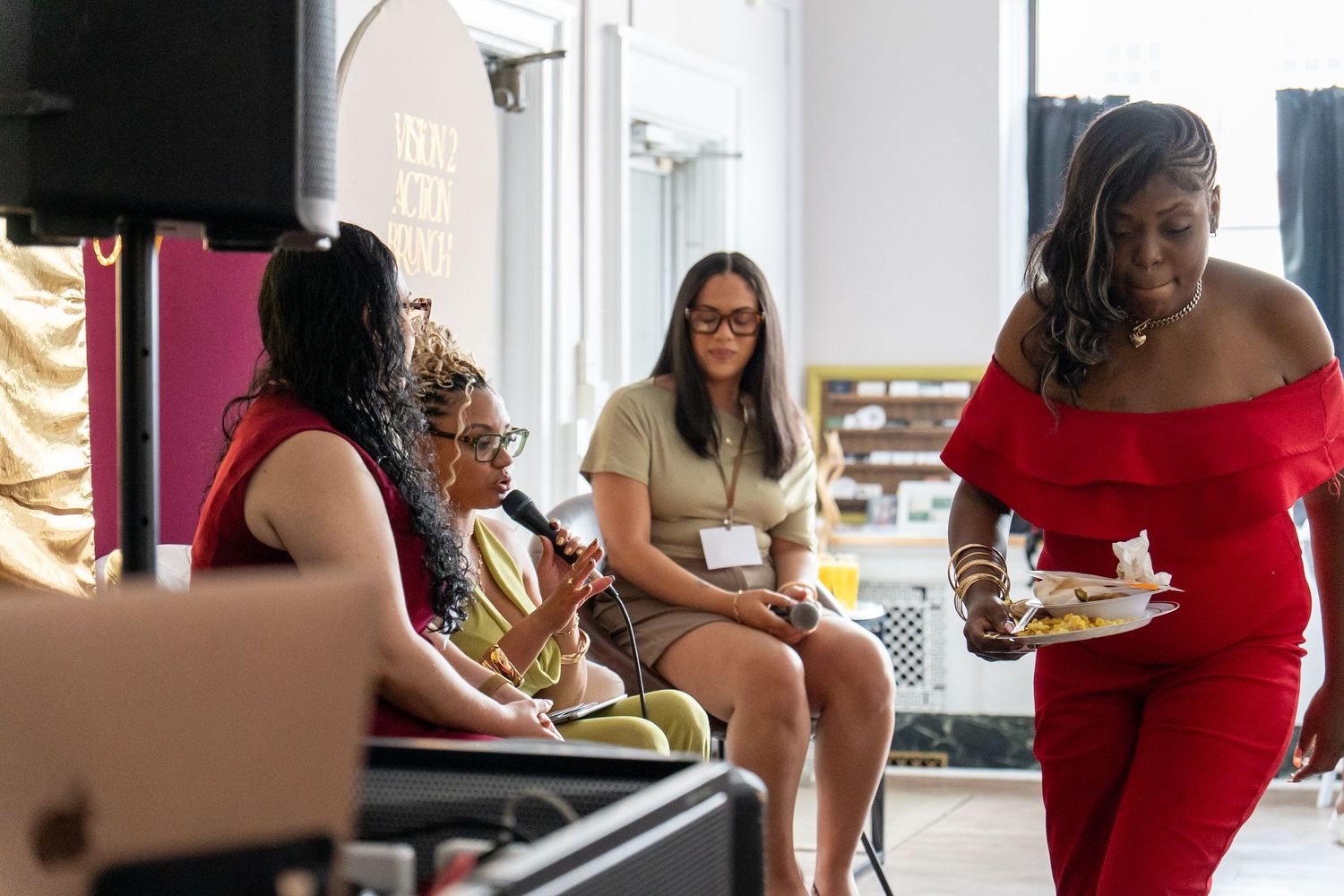 Women at a panel event, one in red dress holding food, others seated with microphone in bright venue.