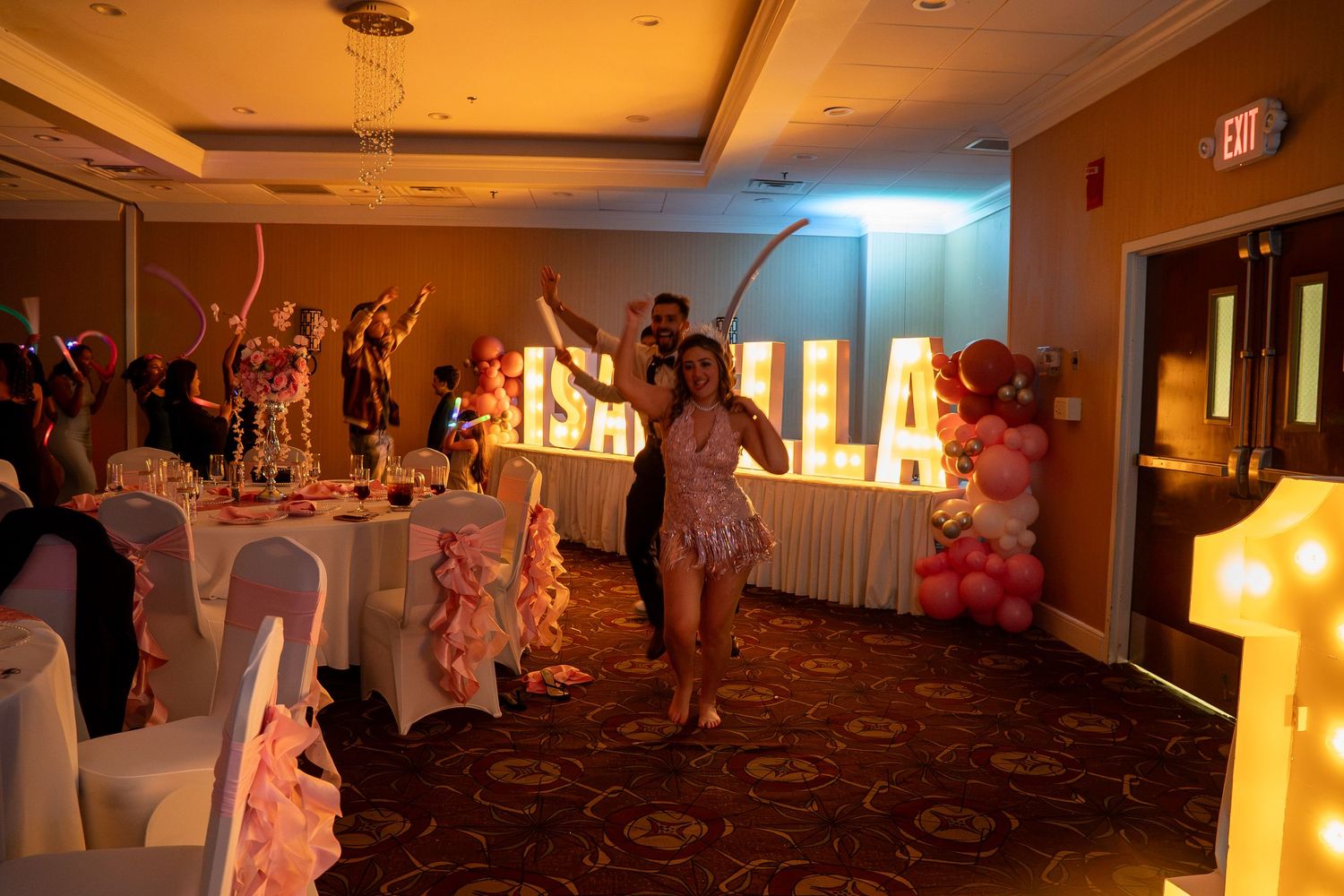 The quinceañera honoree poses near glowing Isabella marquee letters and pink balloon decorations. Sweet 16!