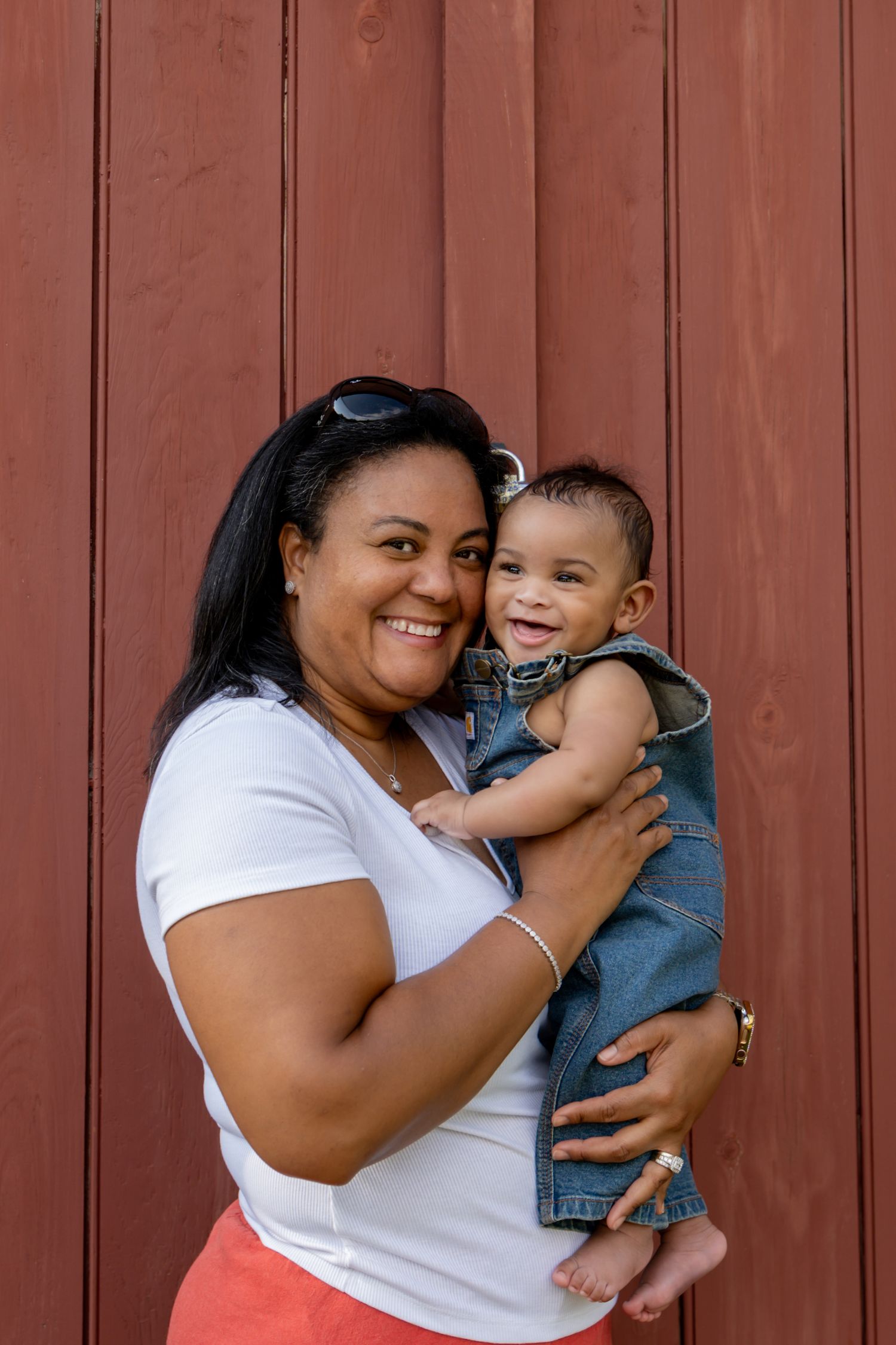 Smiling woman in white shirt holds laughing baby in denim jacket against red wooden wall outdoors. 6 month photoshoot