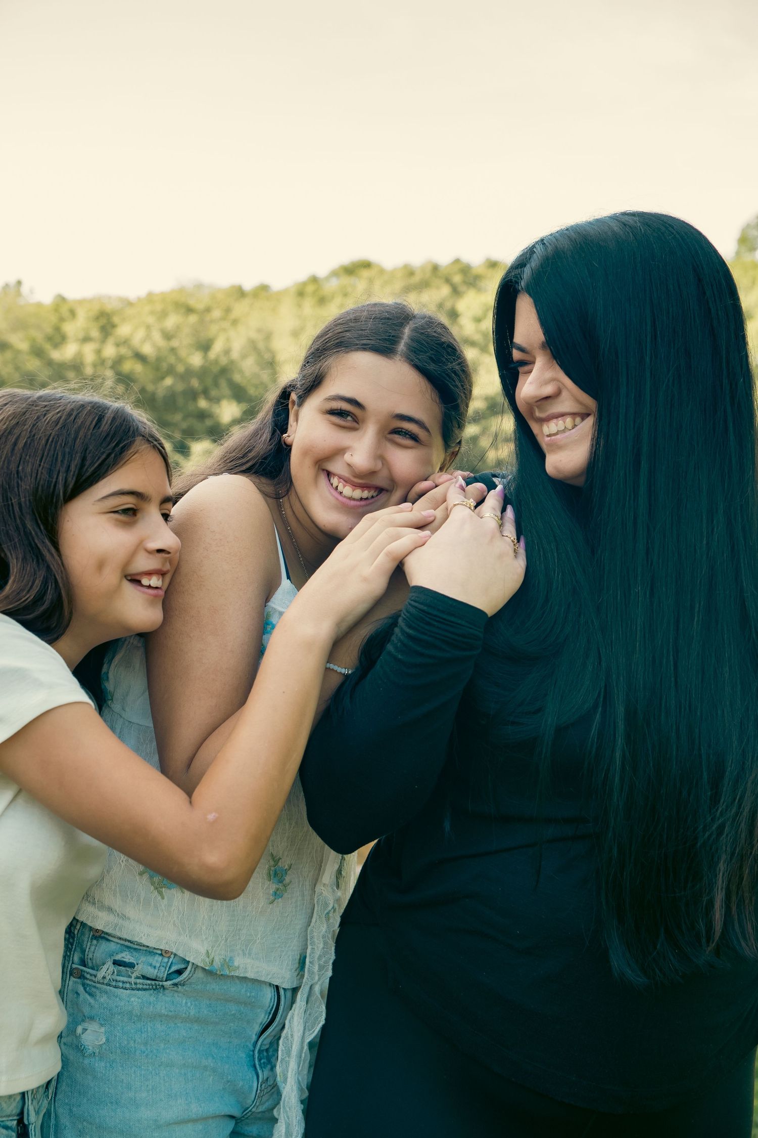 Three smiling women hugging and laughing together outdoors, with a woman with long black hair on the right and green trees behind.
