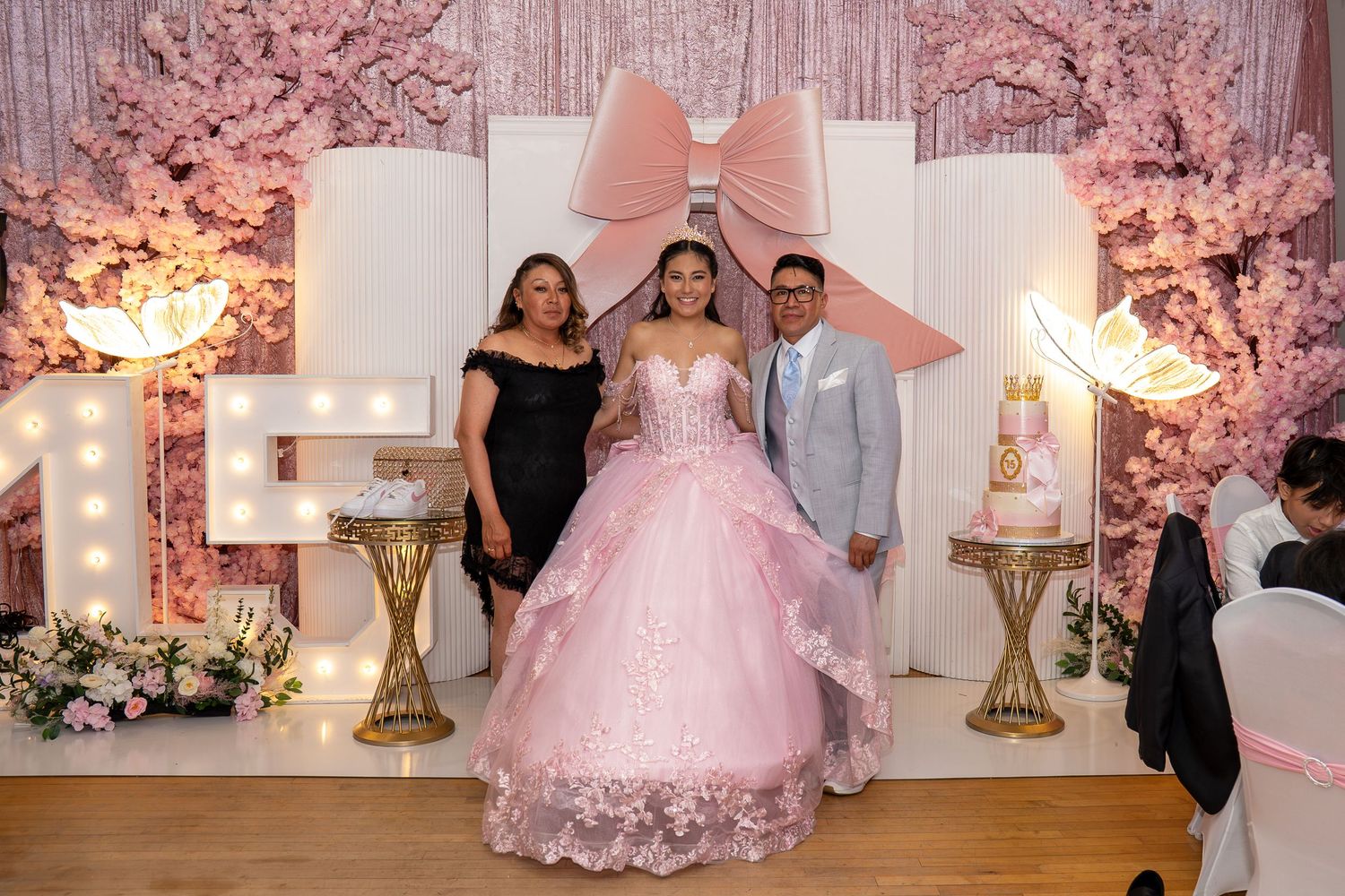 Quinceañera girl in pink gown poses with parents at elegant floral party venue.