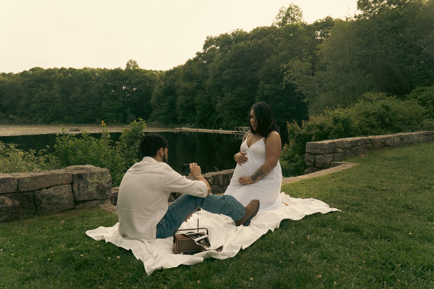 Couple enjoying a romantic outdoor picnic on a white blanket by a serene lake surrounded by lush green trees.