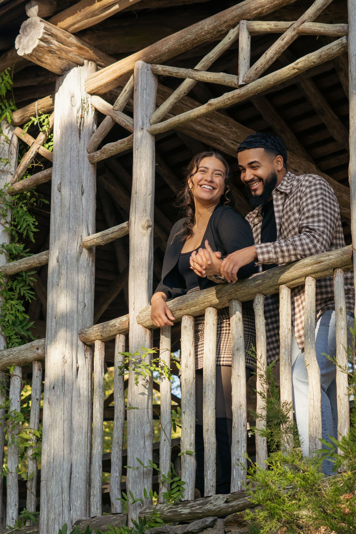 Smiling interracial couple leaning on a rustic wooden railing in a log structure surrounded by greenery.