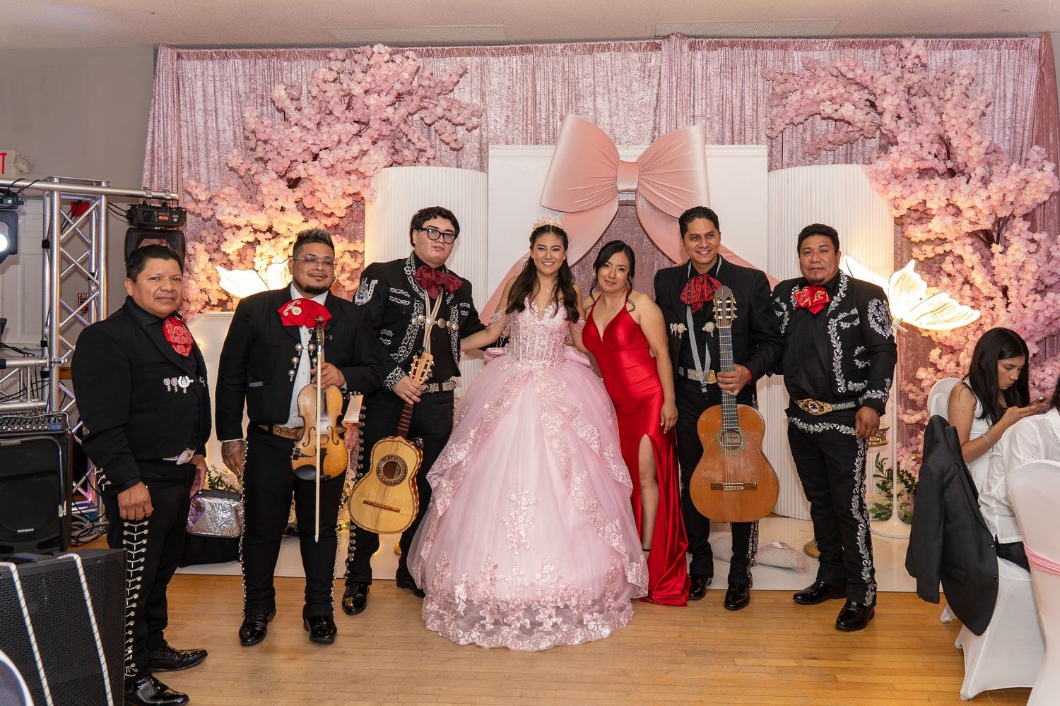 A quinceañera girl in pink gown poses with mariachi band members at an elegant floral decorated celebration.