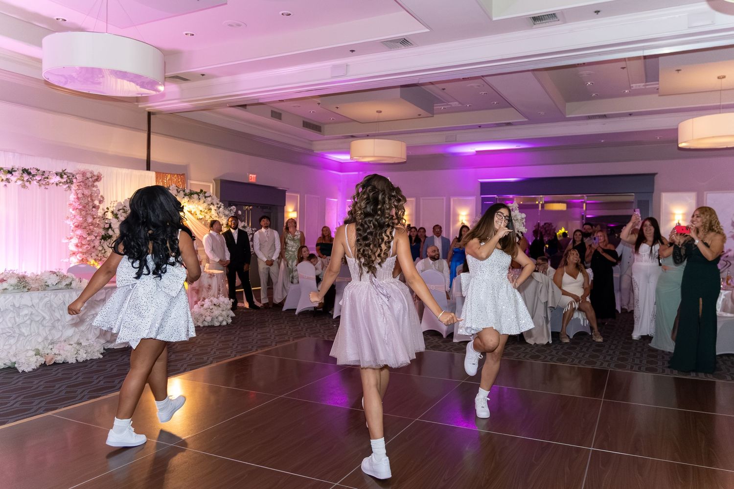 Girls in white dresses perform a choreographed dance at a quinceañera celebration in a decorated ballroom. Sweet 16/Quinceañera
