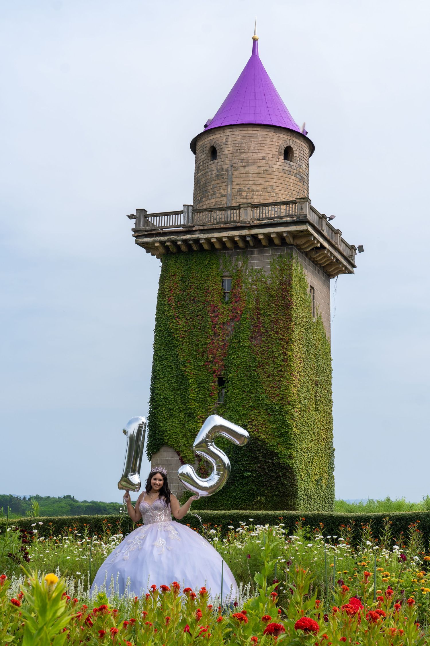 Quinceañera girl in white ball gown holding silver 15 balloons near ivy-covered tower.