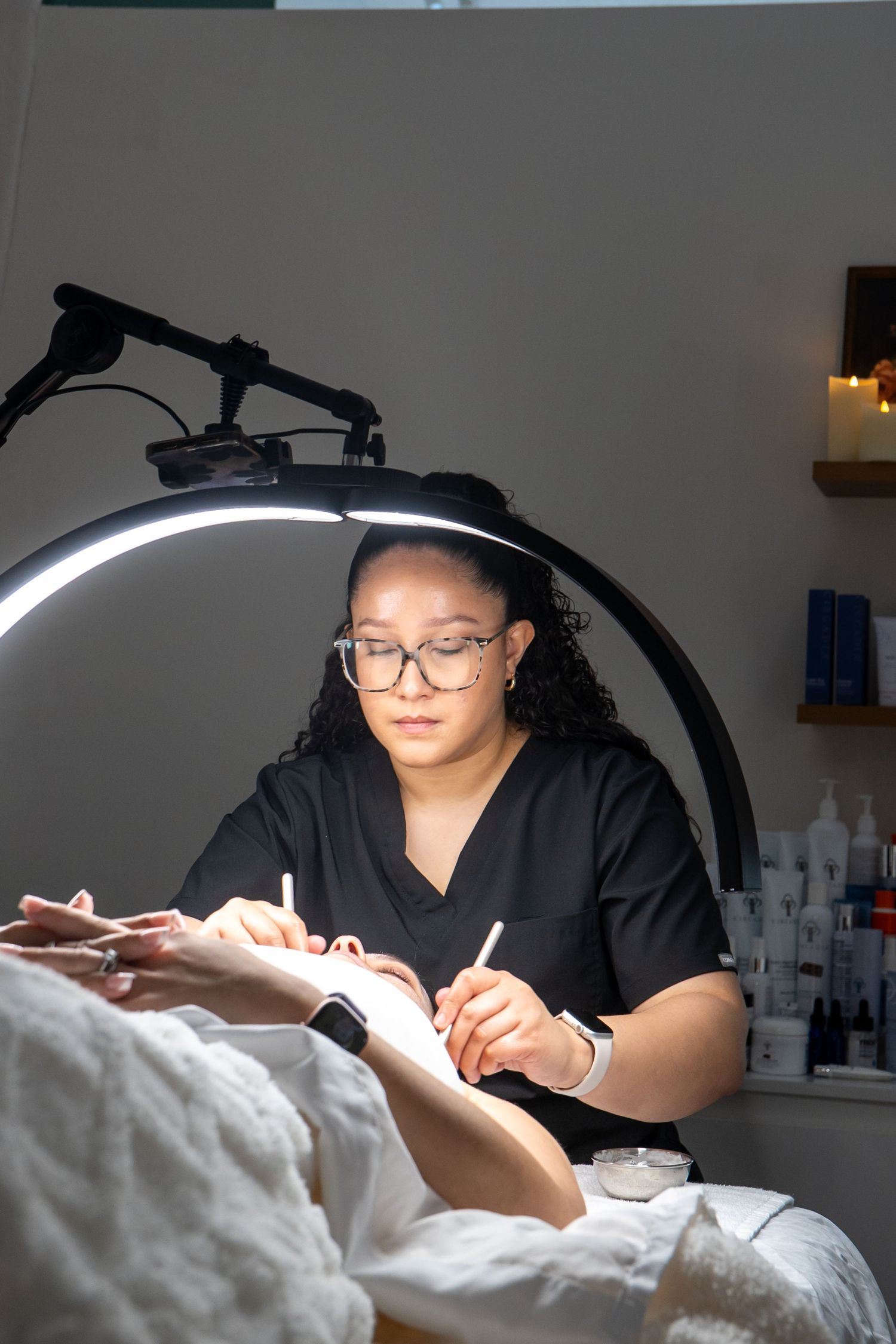 Esthetician in black scrubs applying eyelash extensions under ring light in a professional beauty salon.