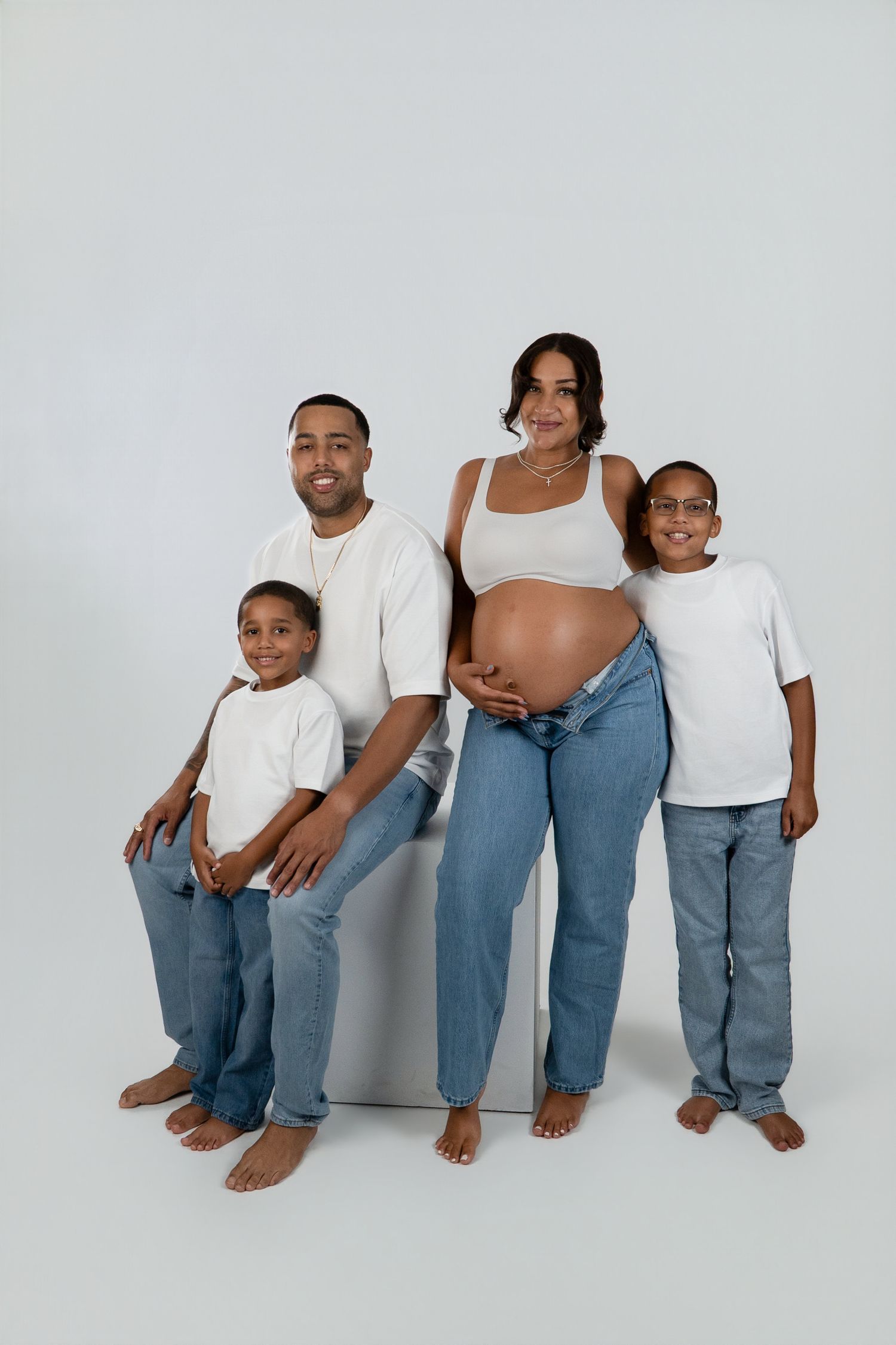 Pregnant mother with bare belly poses with her husband and two sons in white tees and jeans for a maternity family photo.
