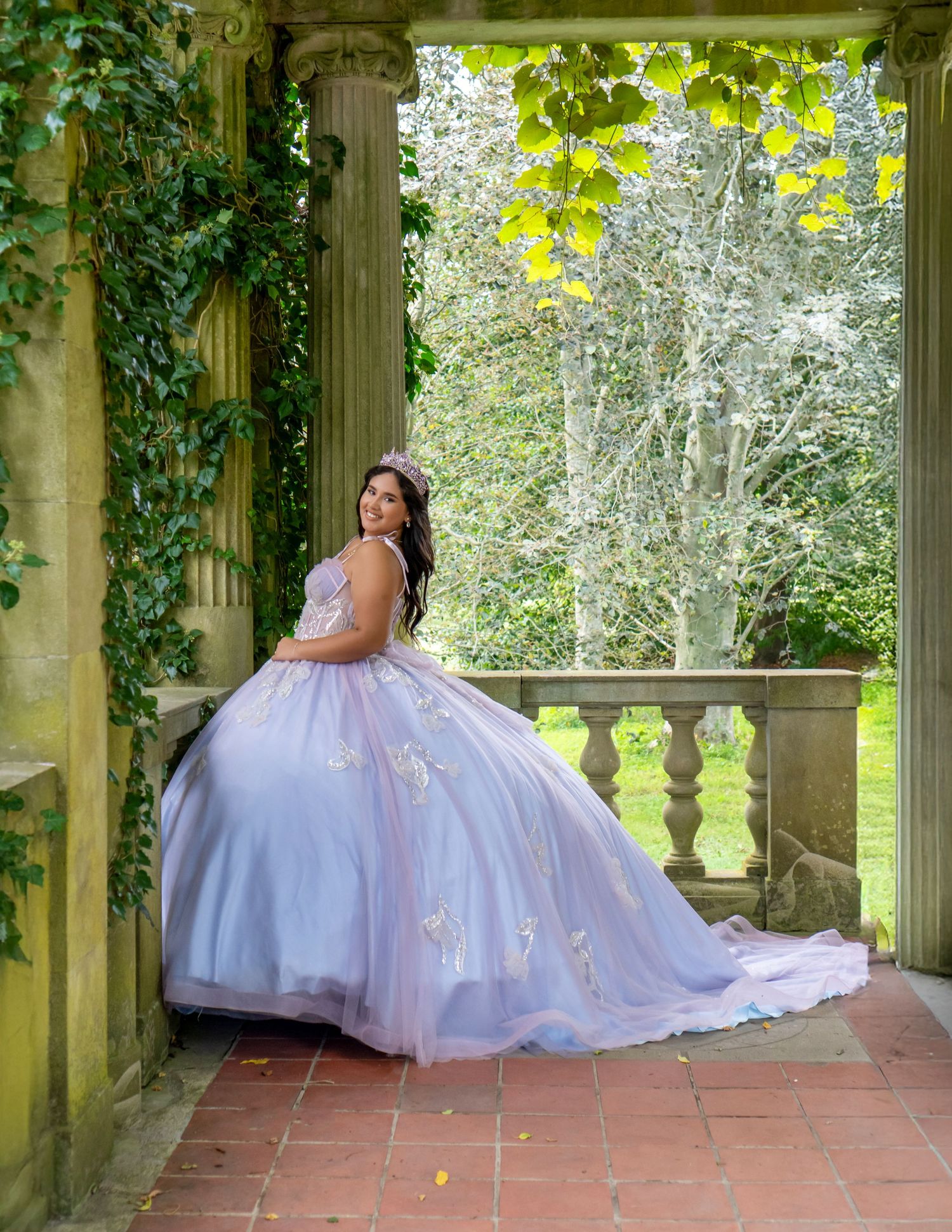 Young woman in lavender quinceañera ball gown posing in ivy-covered stone pavilion.