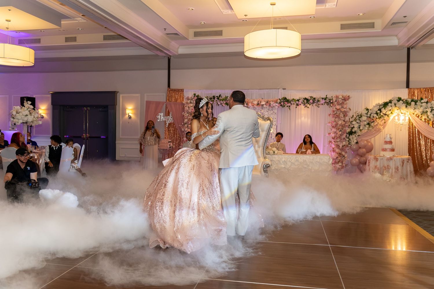 Couple dancing on fog-covered floor at elegant quinceanera reception with pink floral decor and soft purple lighting. Sweet 16/Quinceañera