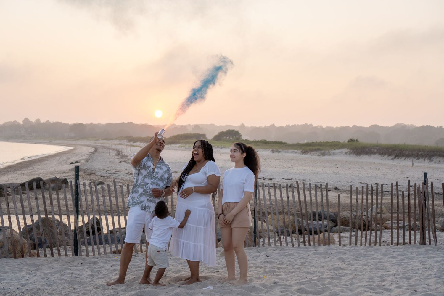 Family in white outfits holds blue smoke flare on beach during golden sunset.