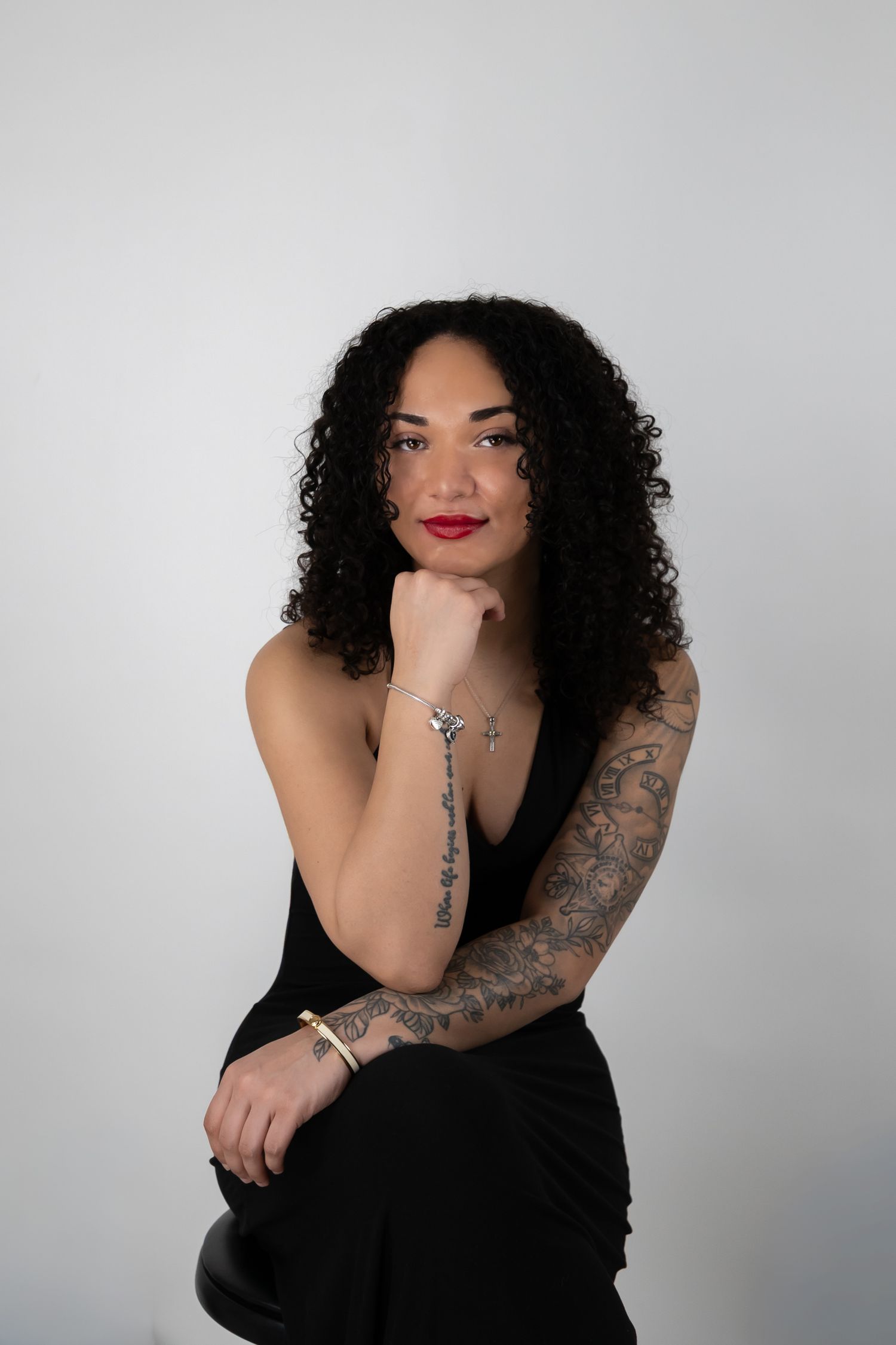 Tattooed woman with curly hair in black dress posing on stool with red lips.