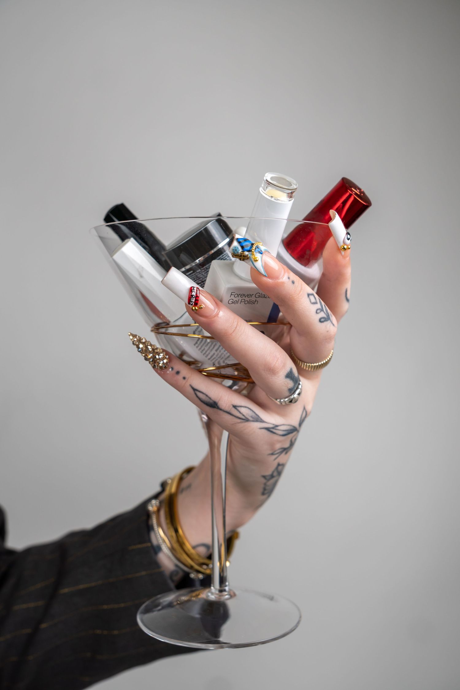 Tattooed hand with long acrylic nails holds a martini glass filled with nail polish bottles.