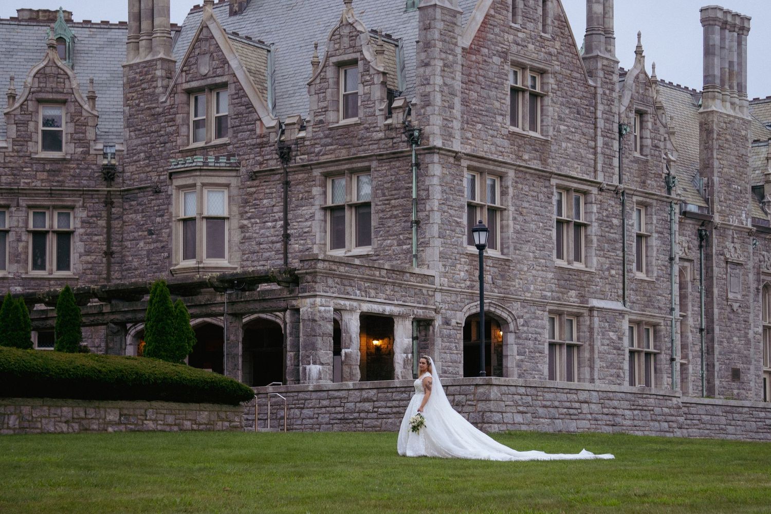 Elegant bride in long white gown posing before grand stone castle venue.