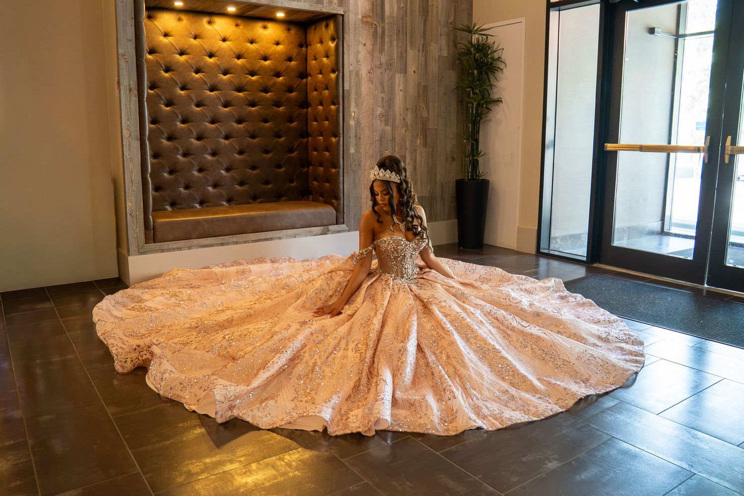 A woman in an ornate gold ballgown with a long train poses in an elegant lobby for a quinceañera or wedding. Sweet 16/Quinceañera