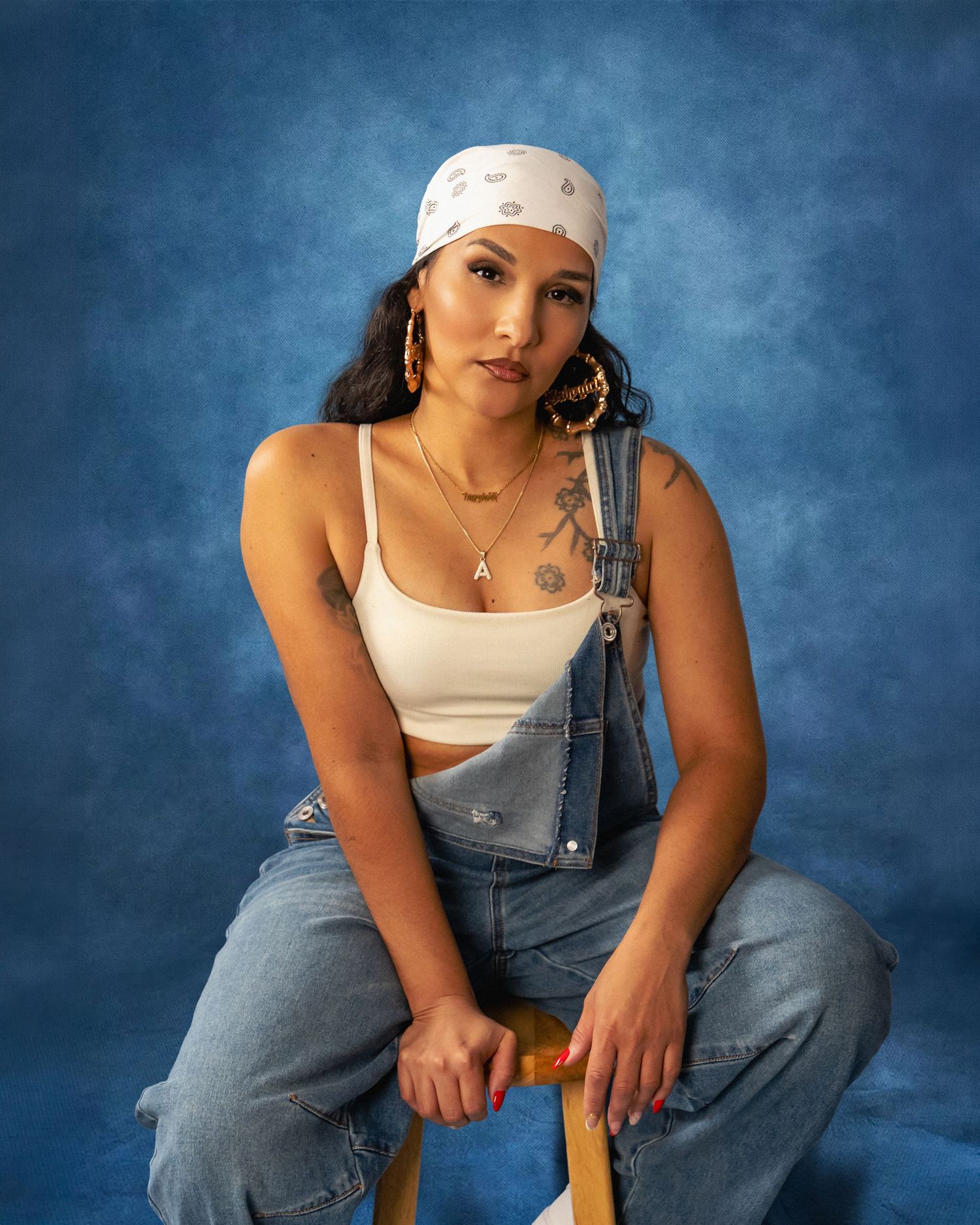 Woman in a white bandana, cropped tank top and denim overalls sits on a wooden stool against a blue studio backdrop.