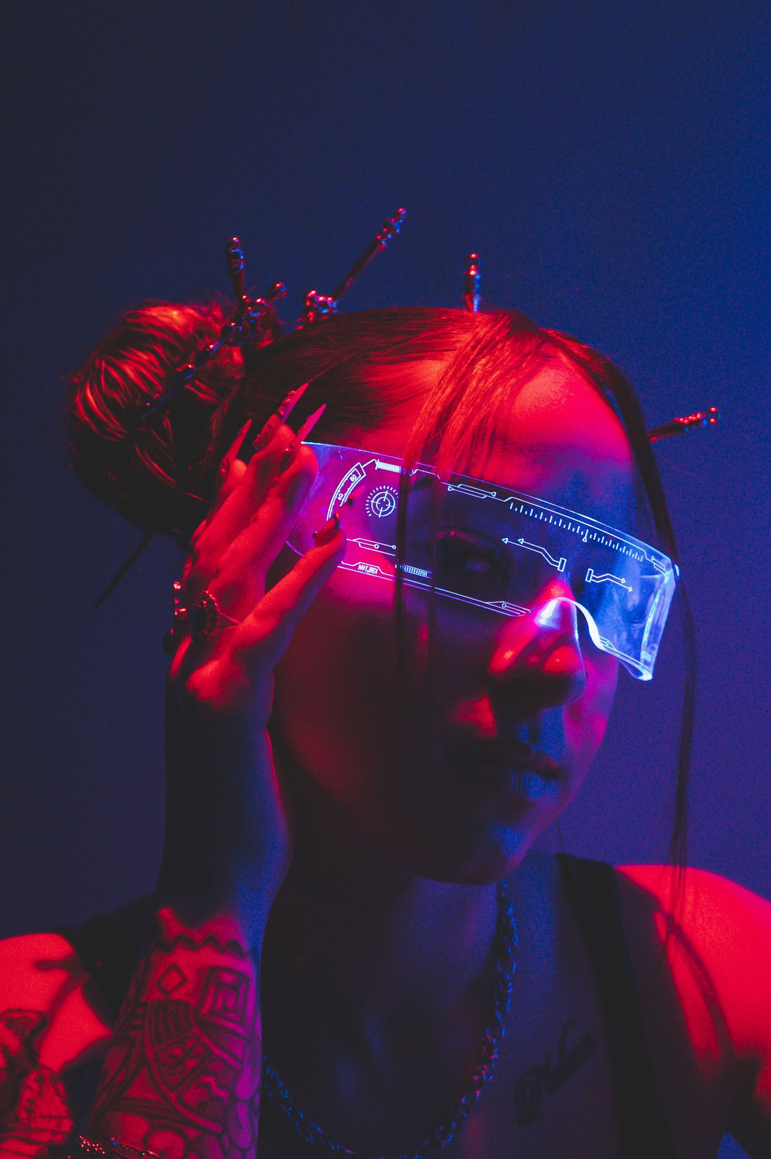 Woman with cyberpunk LED visor glasses and spiked hair accessory posed under dramatic red and blue neon lighting.