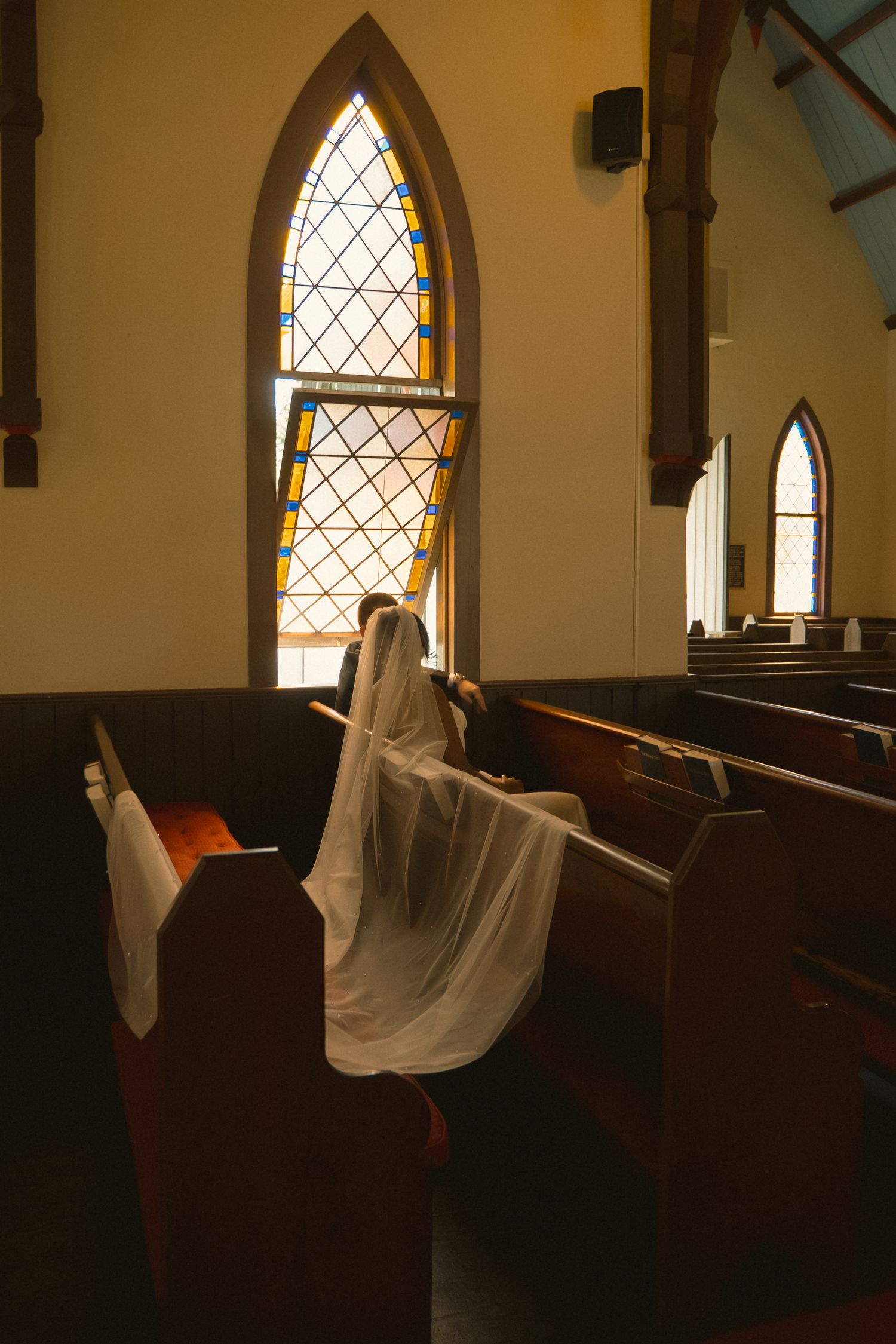 Ghost costume figure sitting in wooden church pew near ornate stained glass windows in historic chapel.